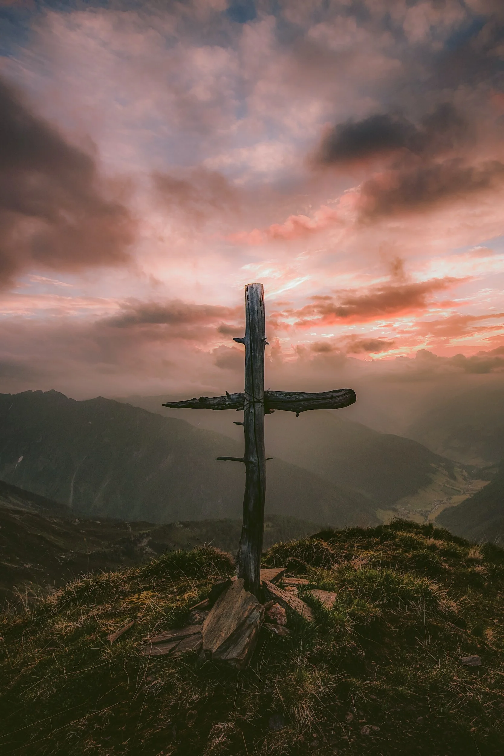 A weathered wooden cross standing on a grassy hill with mountains and a colorful cloudy sky at sunset.