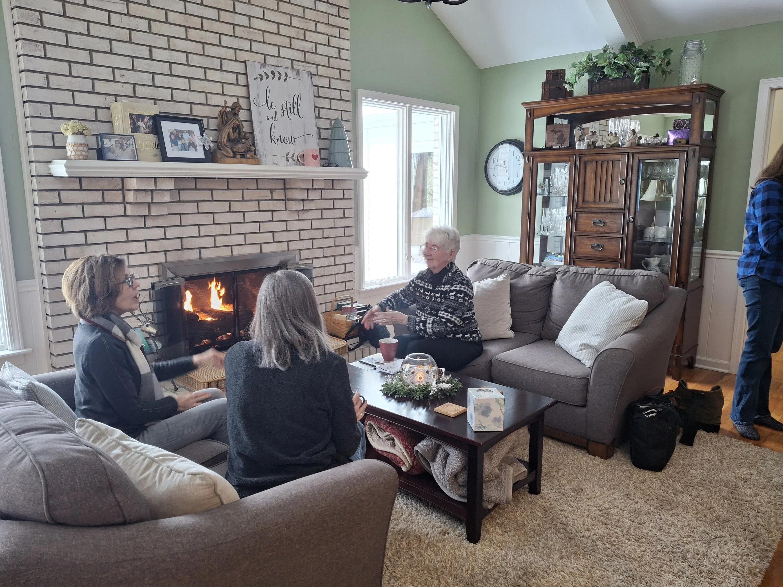 Three women sitting in a living room near a fireplace with a fire, engaging in conversation. One woman is sitting on a couch, another on an armchair, and the third on a nearby chair. There is a decorated table with a candle and greenery, and shelves 