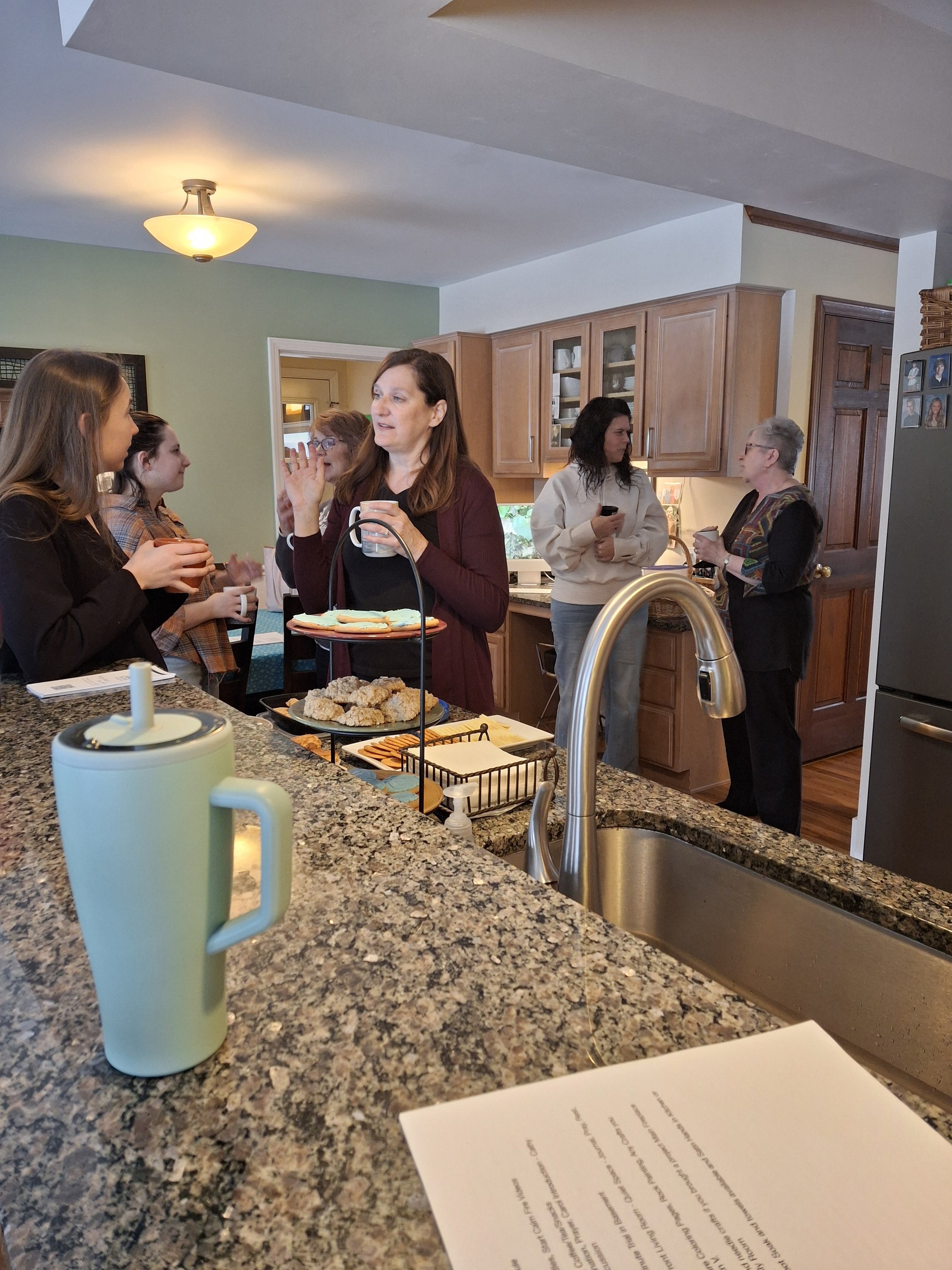 People gathered in a kitchen having conversations; a woman in a maroon top is holding a mug and talking to a young girl, others are standing and chatting near kitchen cabinets and a refrigerator.