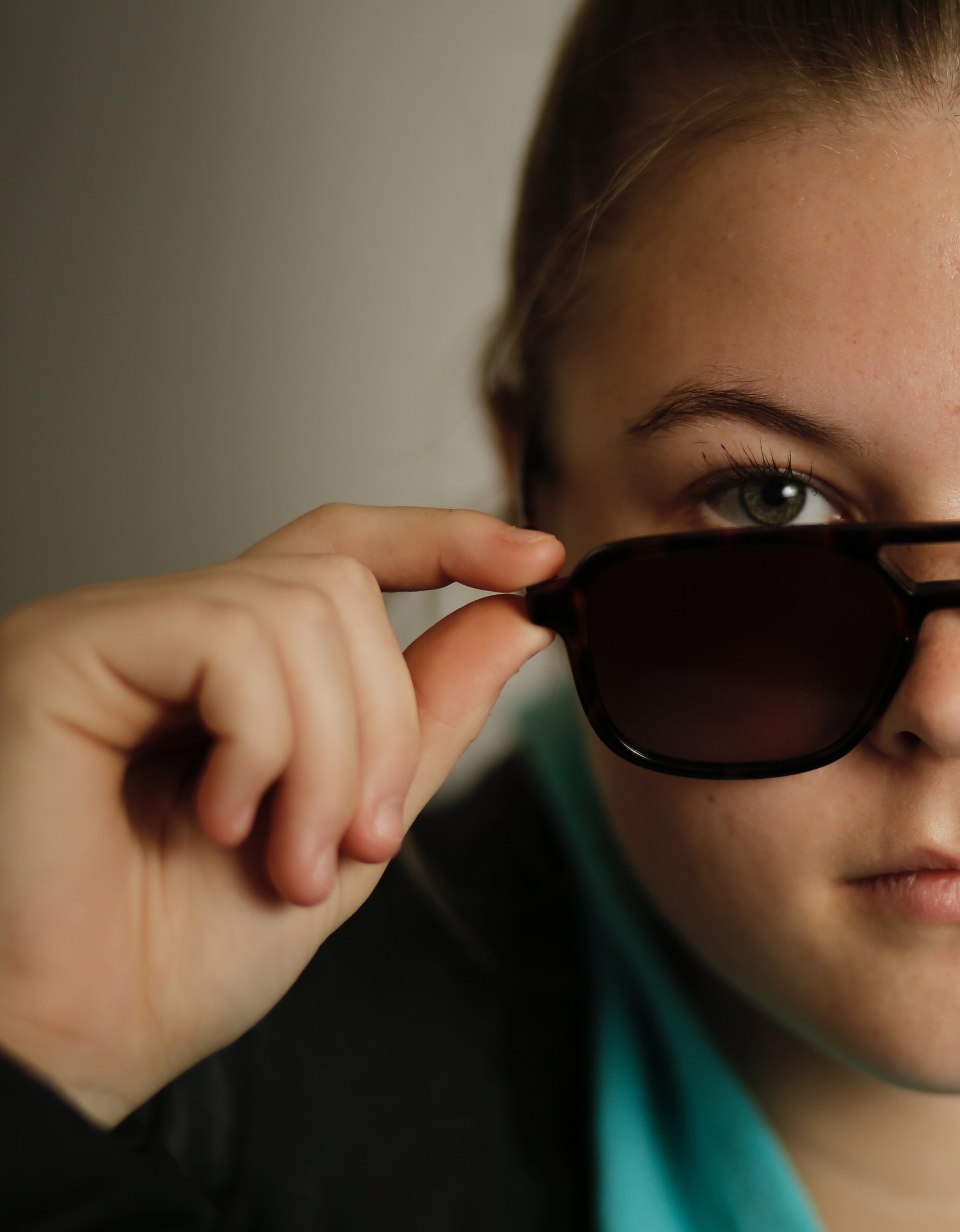 A person with green eyes is holding sunglasses near their face, looking over the top of them.