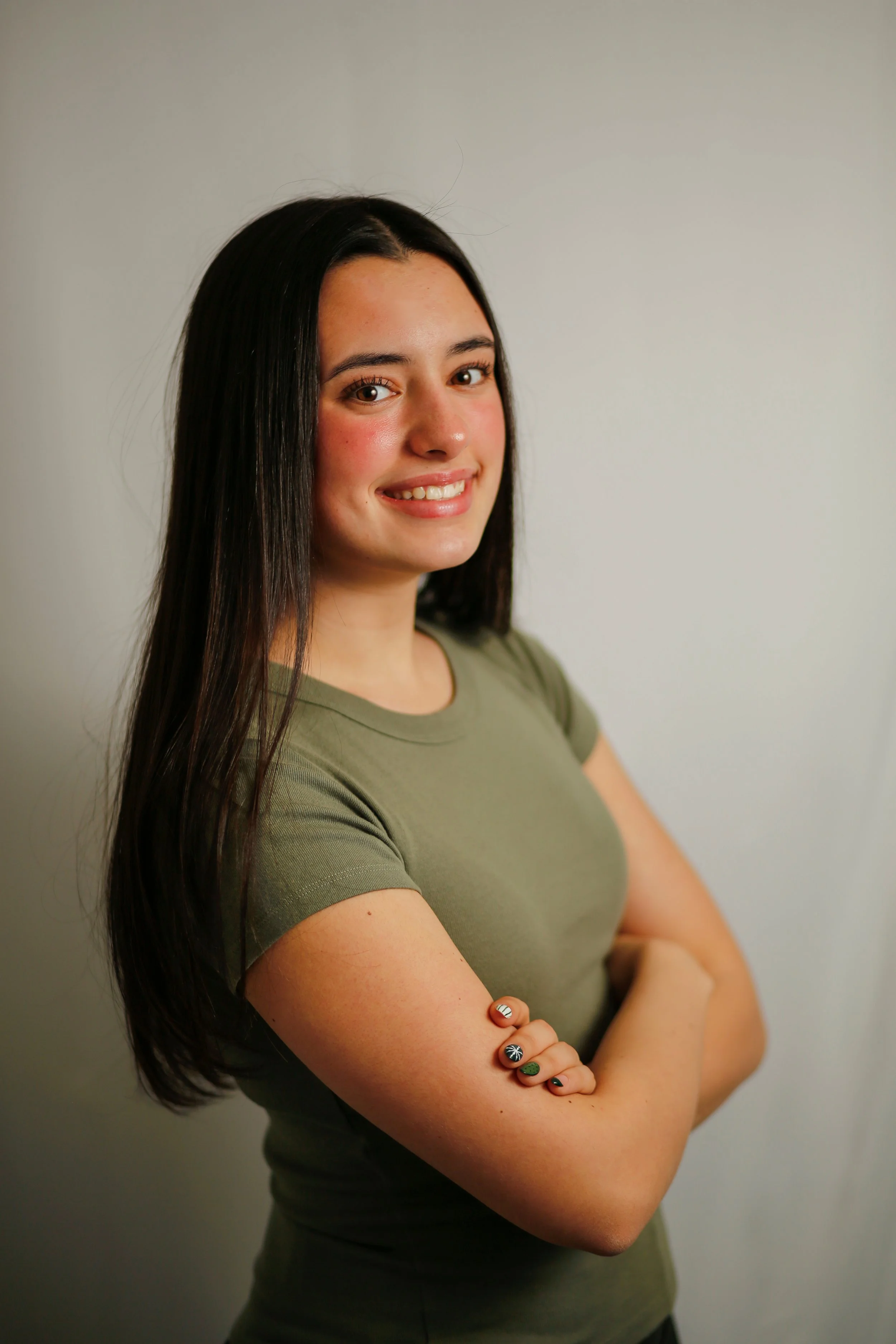 Young woman with long dark hair smiling, wearing a green t-shirt, arms crossed, standing against a white wall.