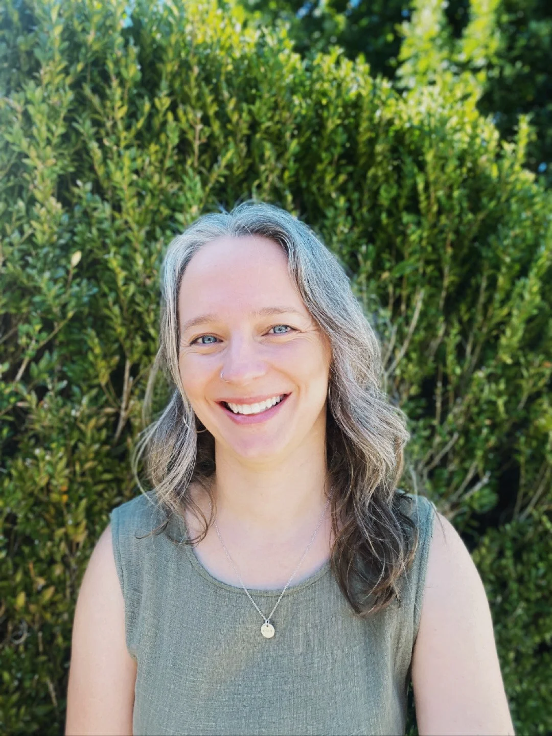 A woman with shoulder-length gray hair, wearing a gray sleeveless top and a silver necklace with a small round pendant, smiling outdoors against a background of green bushes.