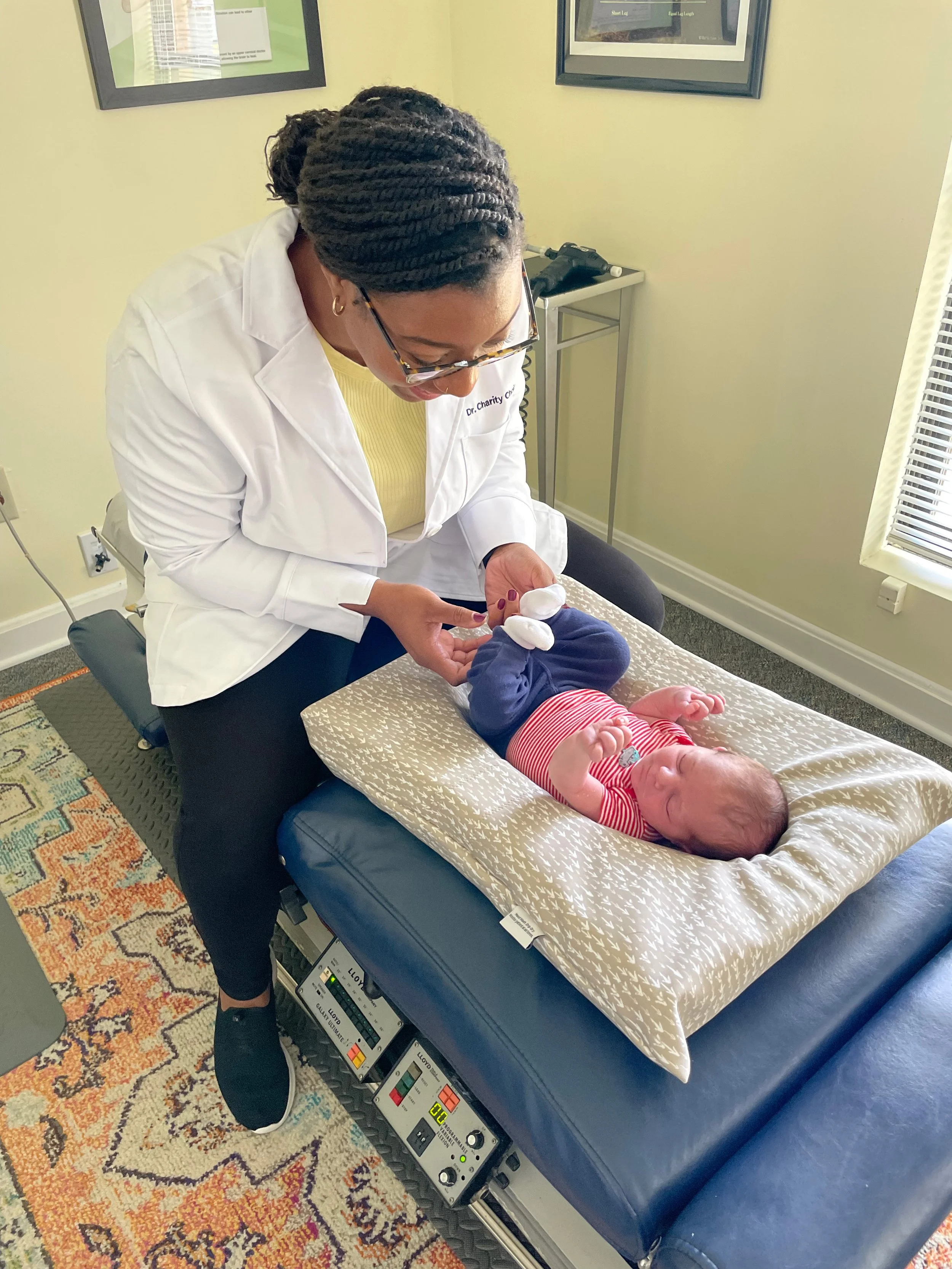 Dr. Choice, owner of Radiate Chiropractic, likely a doctor, is examining a baby on an examination table. The baby is lying on a cushioned pad, wearing a red and white striped shirt and blue pants, and appears to be relaxed.