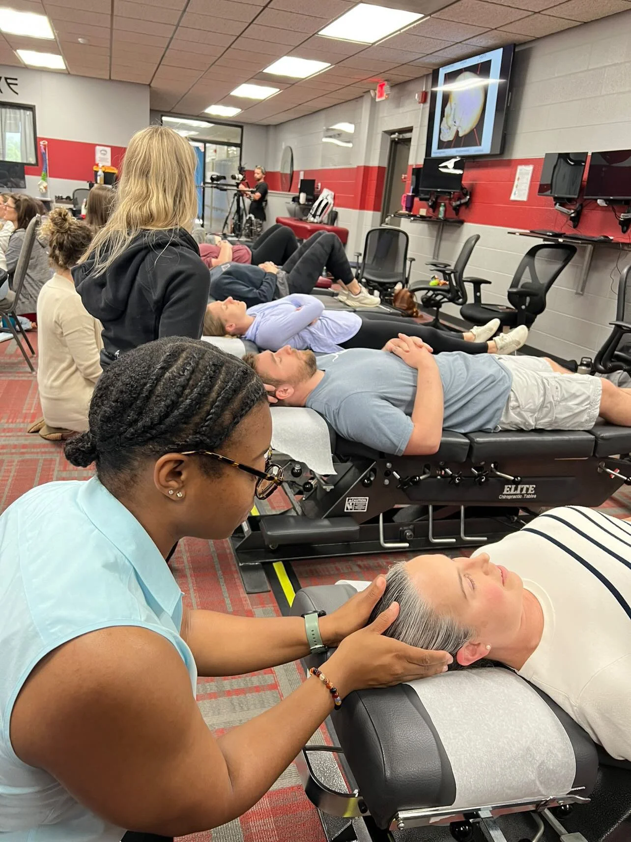 A group of people lying on chiropractic tables receiving neck adjustments from Dr. Choice, owner of Radiate Chiropractic in a classroom or seminar setting equipped with multiple screens and chairs.