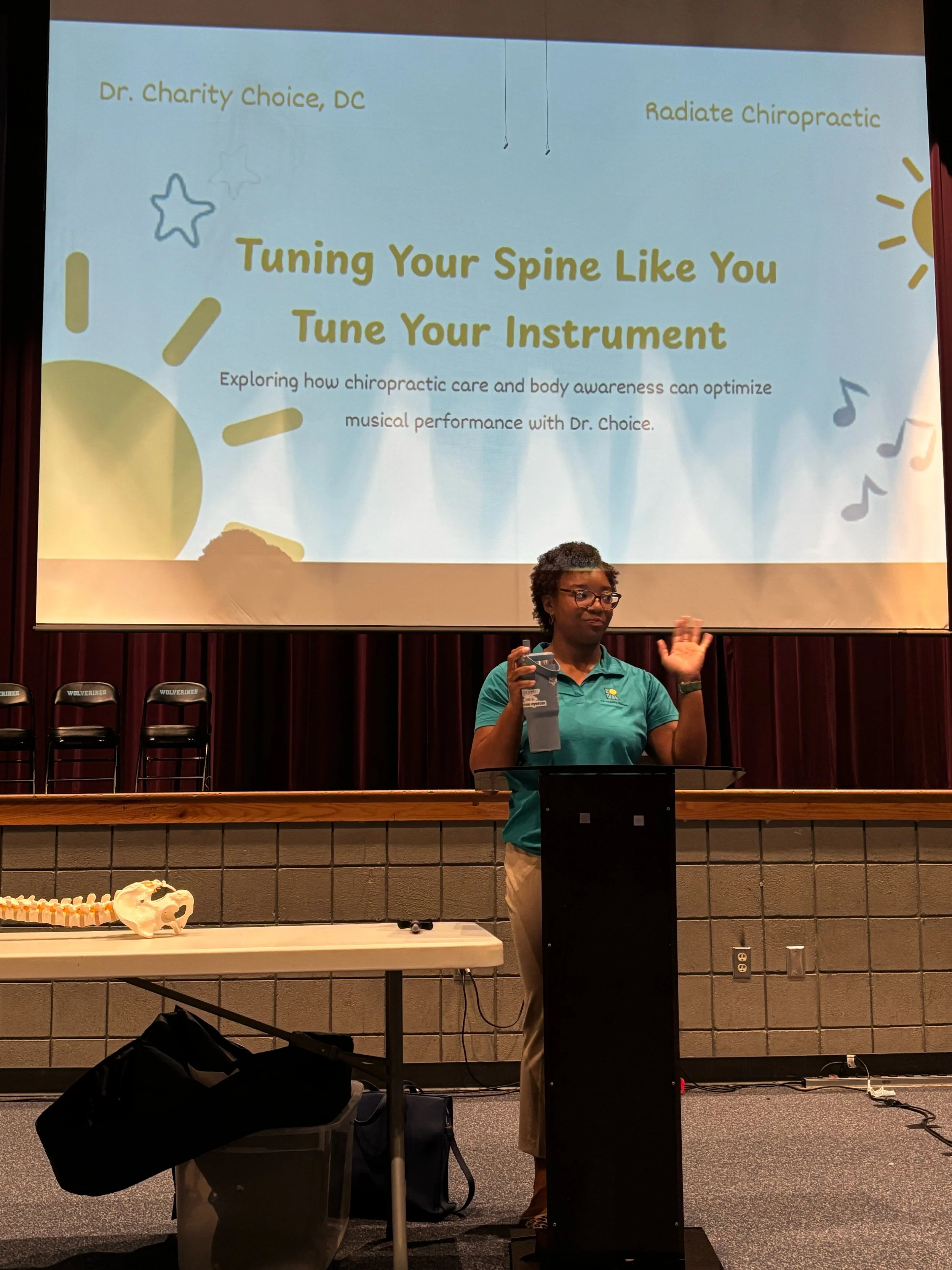 Dr. Choice, owner of Radiate Chiropractic a podium in front of a presentation slide titled 'Tuning Your Spine Like You Tune Your Instrument,' with the subtitle describing chiropractic care and body awareness.