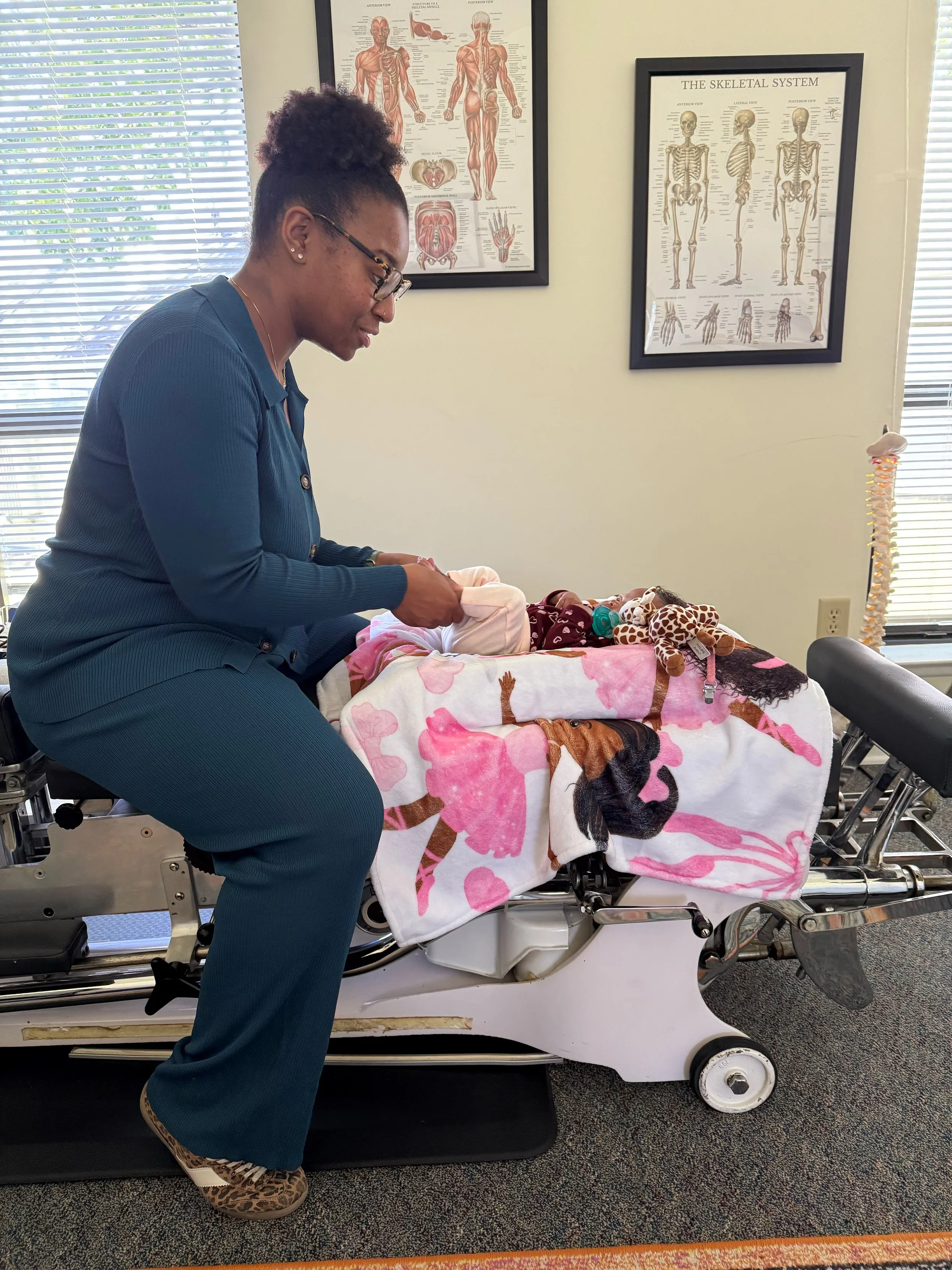 Dr. Choice, owner of Radiate Chiropractic adjusting or caring for a doll on a hospital gurney covered with a pink blanket featuring a horse design.