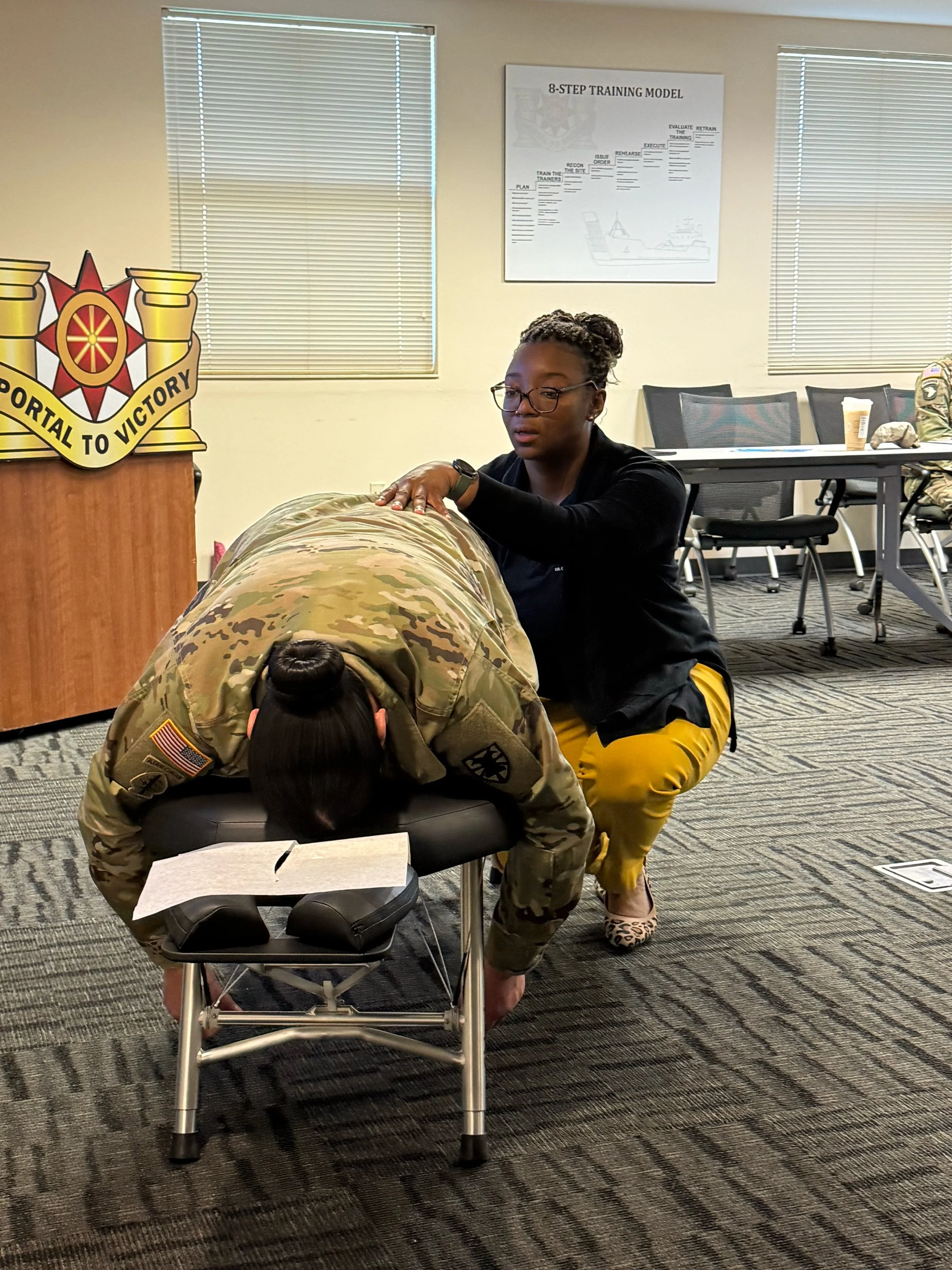 A person in military uniform receiving a neck chiropractic care from Dr. Choice, owner of Radiate Chiropractic in a training room with seating and a poster on the wall titled '8-STEP TRAINING MODEL'.