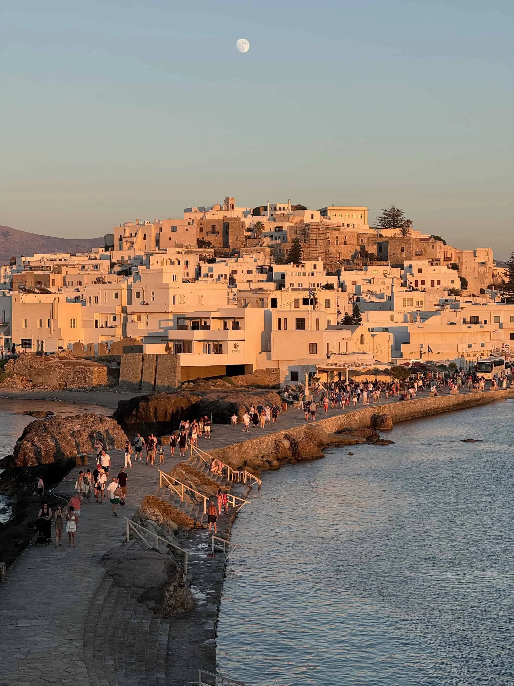 Sunset view of Chora Naxos from the Temple of Apollo, with golden light over the old town and visitors walking along the causeway — a must-see highlight when planning what to do in Naxos.