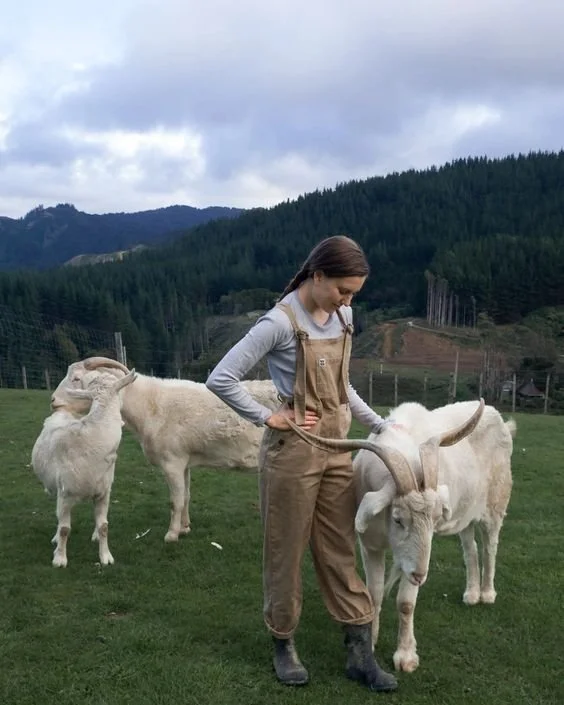 girl in brown overalls tending to goats with horns in pasture