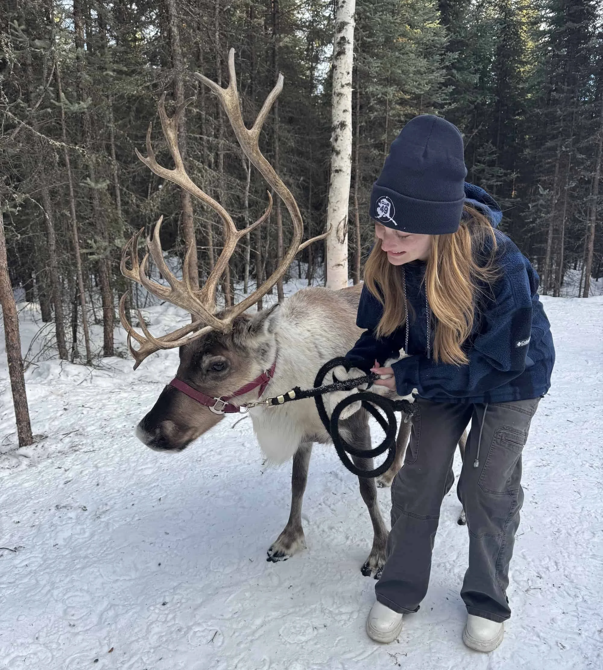 Woman in a beanie and fleece jacket standing next to a reindeer in snowy Fairbanks forest, offering a real-life example of layering for outdoor activities in the Alaskan winter.
