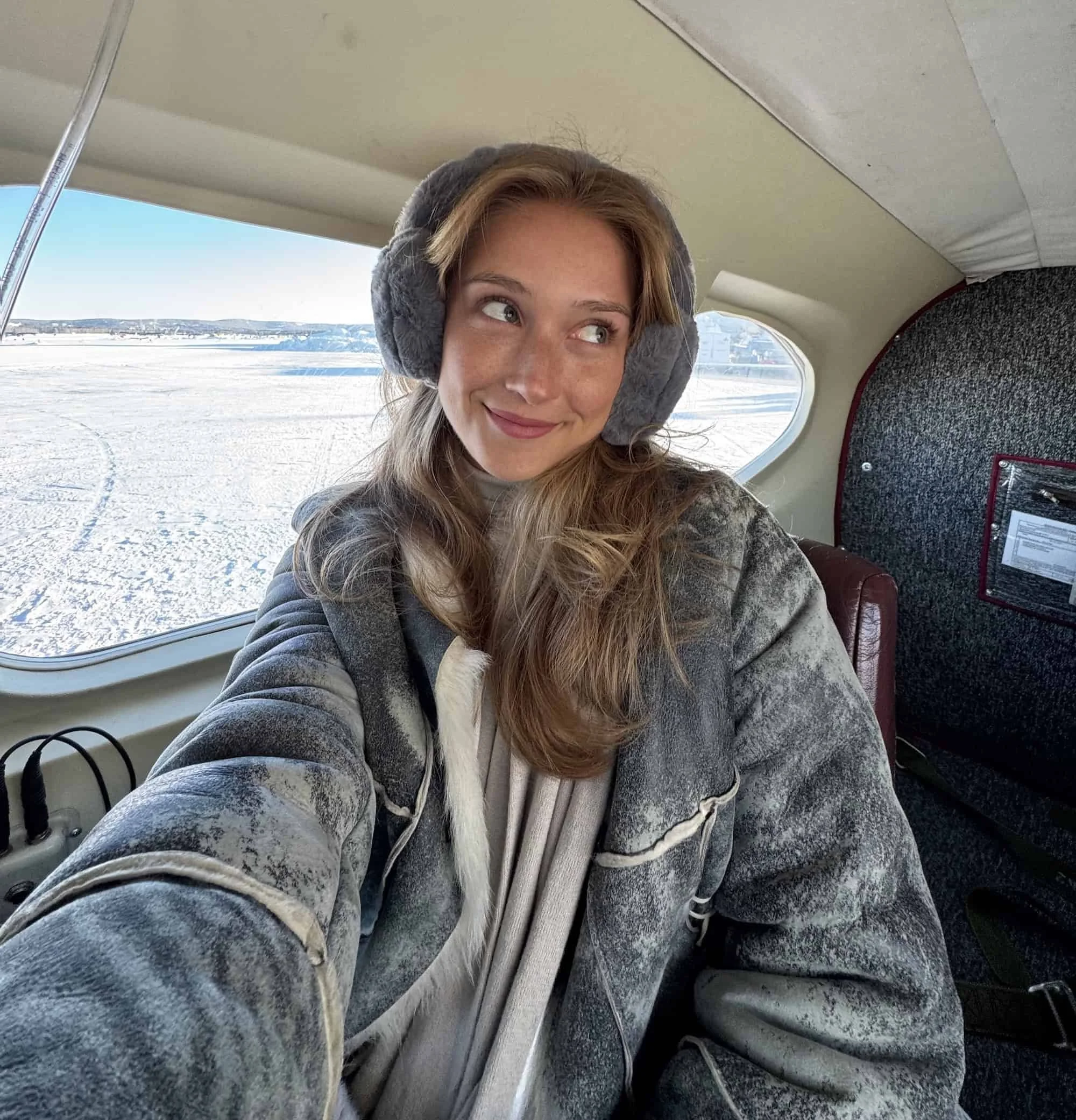 Woman bundled in earmuffs and a shearling-lined coat inside a bush plane over a frozen Alaskan landscape, showcasing how to dress for extreme cold in Alaska’s remote winter settings.