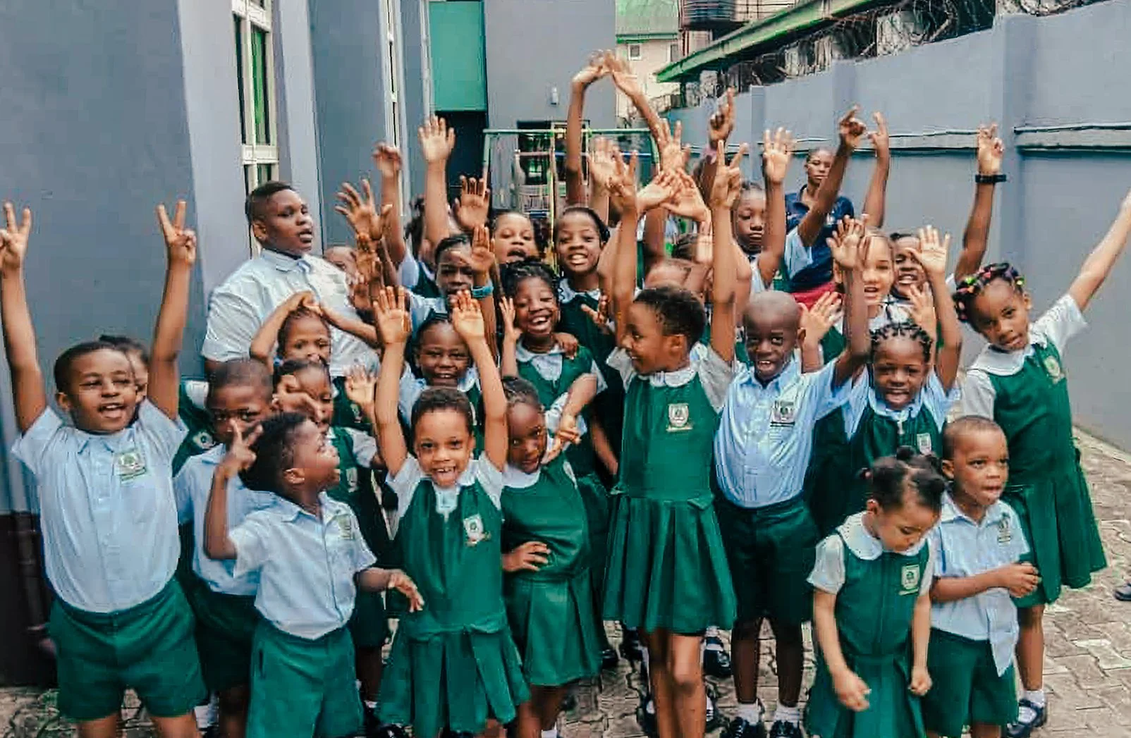 Group of smiling school children in green and white uniforms with some raising their hands, outdoors near gray walls and a green roof canopy.