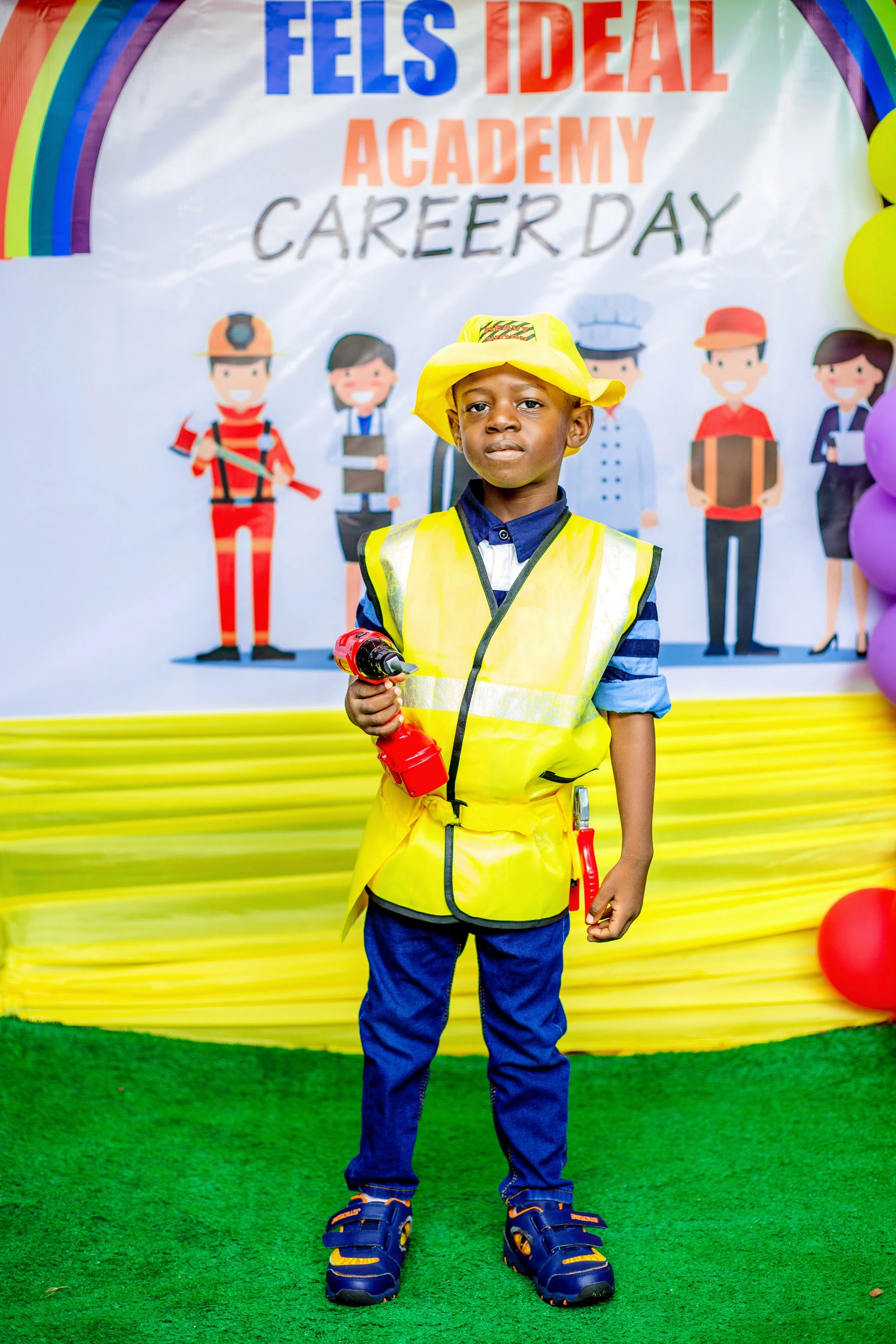 Young boy dressed as a construction worker, wearing a yellow hard hat and a high-visibility safety vest, holding a toy drill, standing in front of a colorful banner at Career Day event.