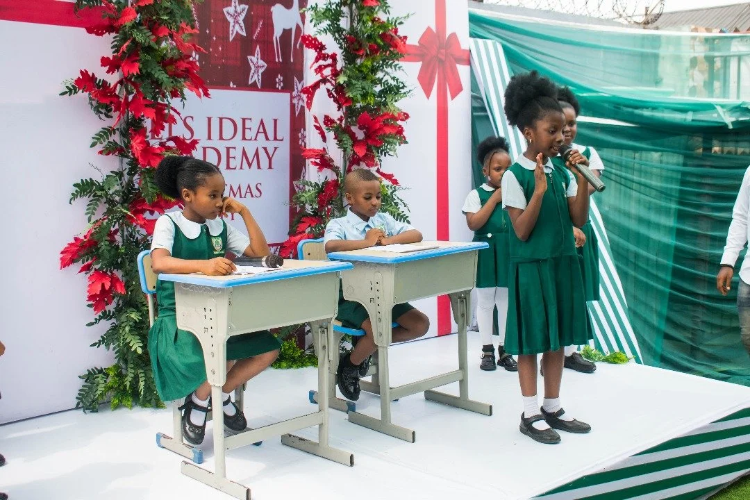 Children on a stage during a Fels Ideal Academy Nursery & Primary school Christmas event, with one girl speaking into a microphone and others sitting or standing, decorated with festive holiday decorations.