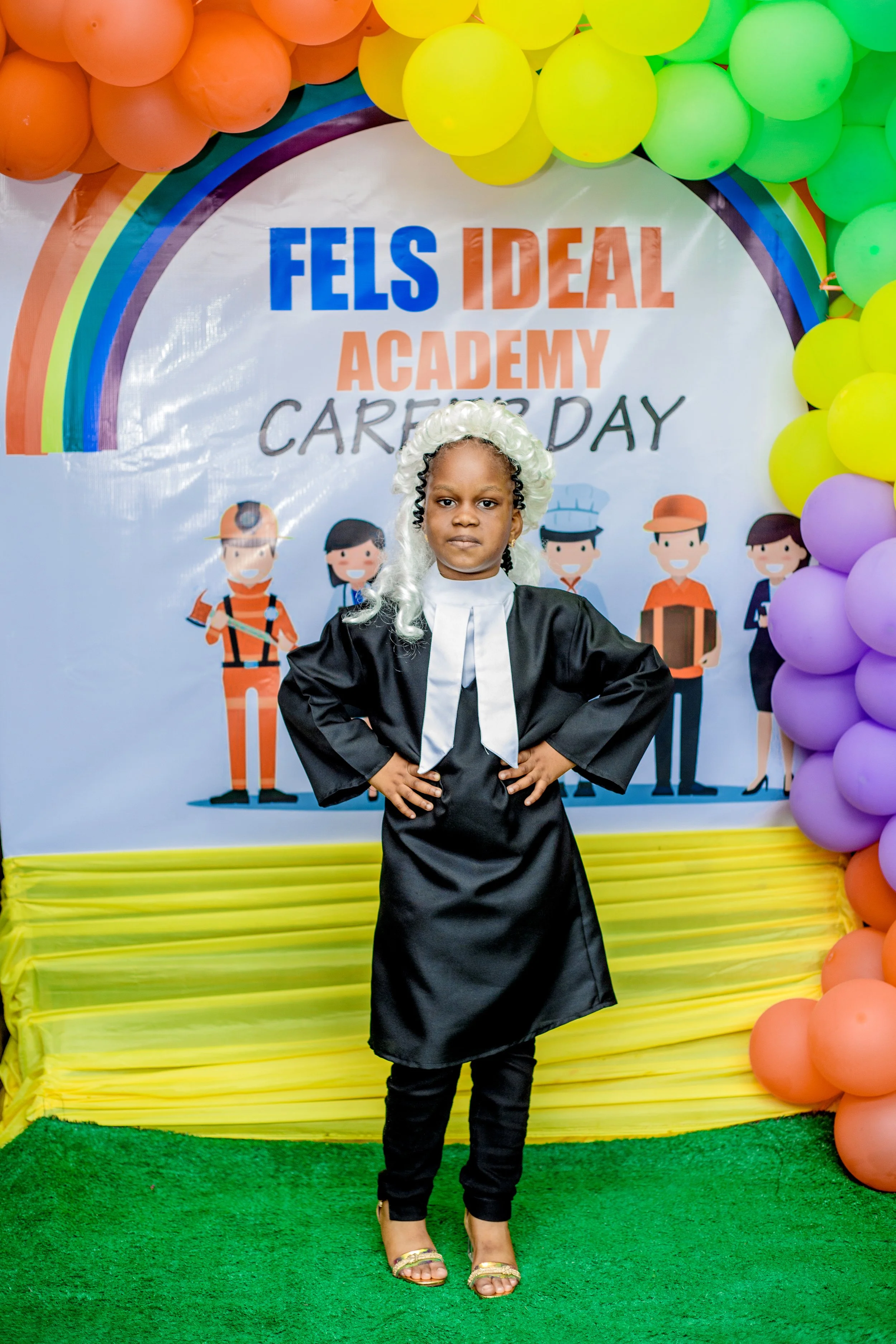 Young girl dressed as a judge, standing confidently with hands on hips, at a career day event with a colorful rainbow-themed backdrop and balloons, celebrating Fels Ideal Academy Career Day.