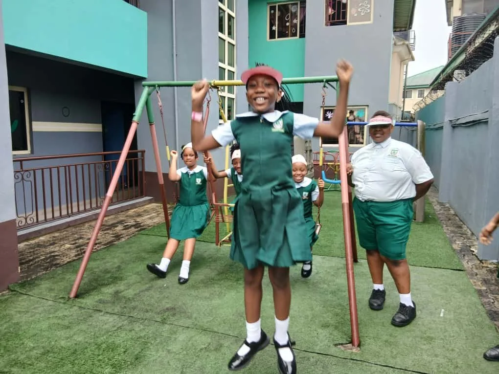 Primary school children in school uniforms playing on a swing set outdoors, with a smiling girl in the foreground hanging on the swing.