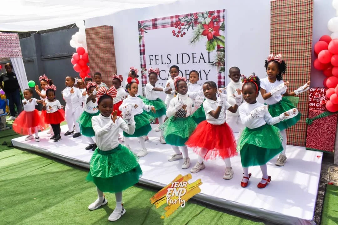 Children performing a dance at a year-end party, dressed in festive red, white, and green outfits, with a decorated backdrop and balloons.