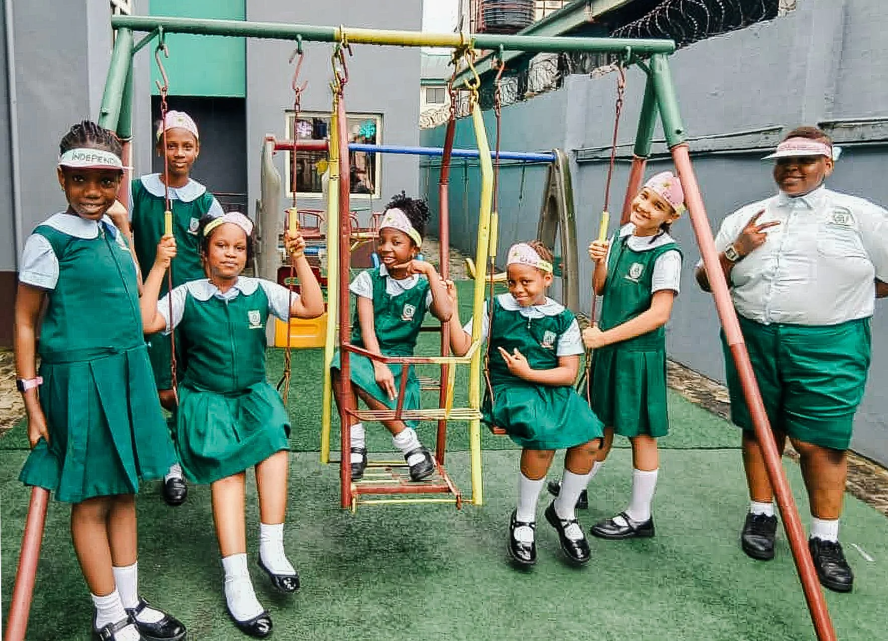 Group of seven young schoolgirls and one woman, likely a teacher, standing and sitting on a playground swing set in school uniform. The girls are wearing green dresses and white shirts with sailor collars, some are holding onto the swing ropes, and the woman is standing on the right side making a peace sign. All are smiling and appear to be enjoying themselves.