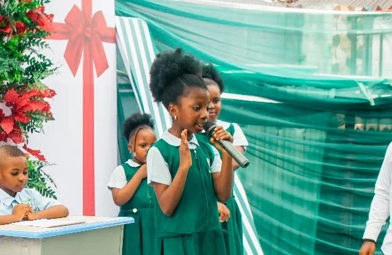 Young girl in green school uniform speaking into a microphone during a school event with other children and Christmas decorations in the background.