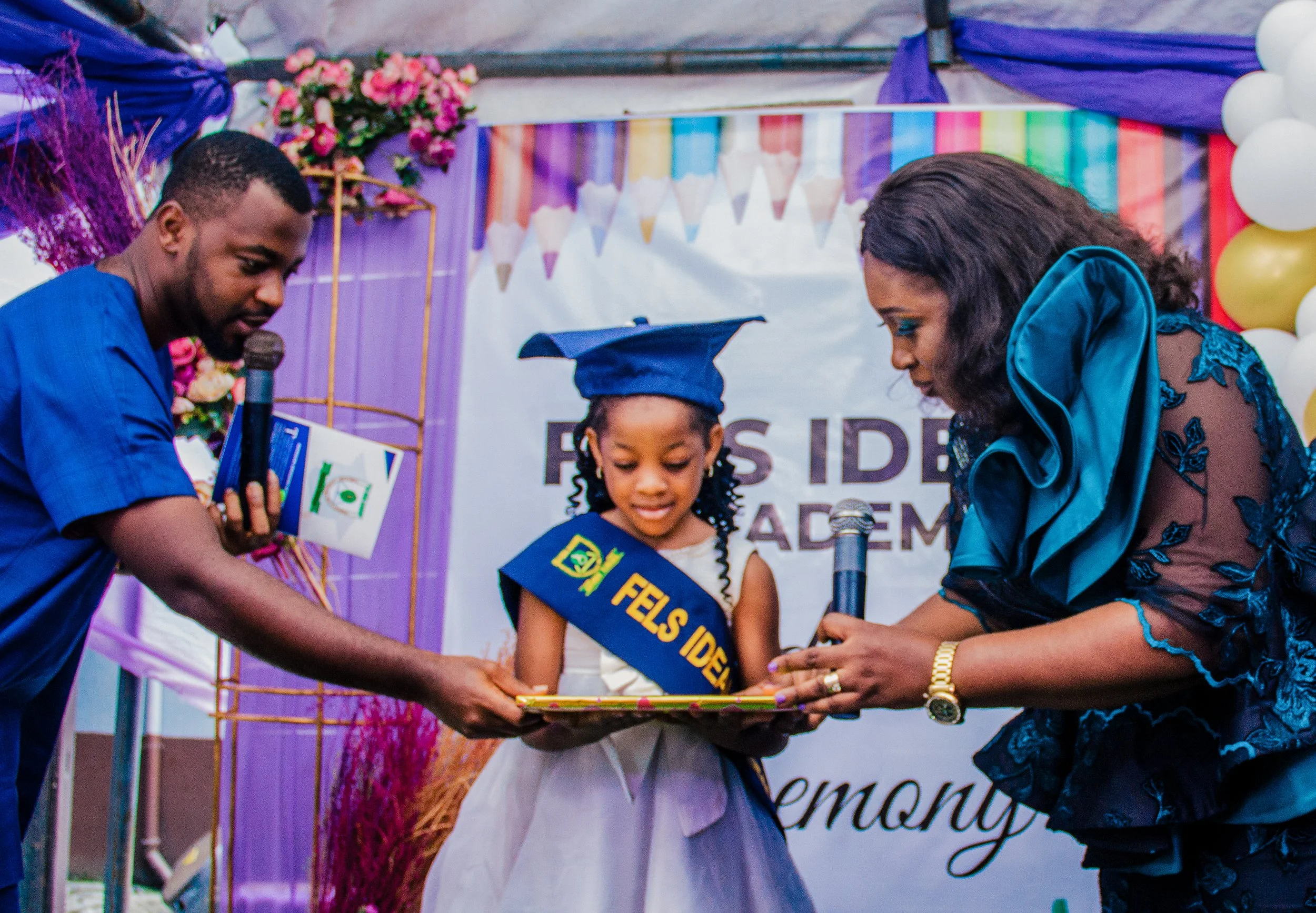 Young girl wearing a blue graduation cap and sash receiving a certificate during a graduation ceremony, with two adults handing her the certificate and holding microphones, decorated with balloons and a colorful backdrop.