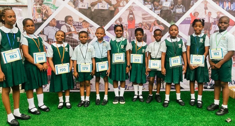 Group of Fels Ideal Academy school children in green and white uniforms standing on green grass, each wearing a badge with a contestant number, for an outdoor competition.