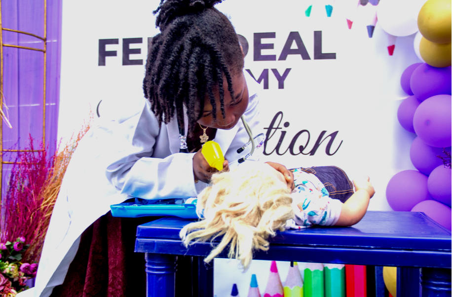 A woman dressed as a doctor examining a child lying on a table at a medical-themed event with balloons and decorations.