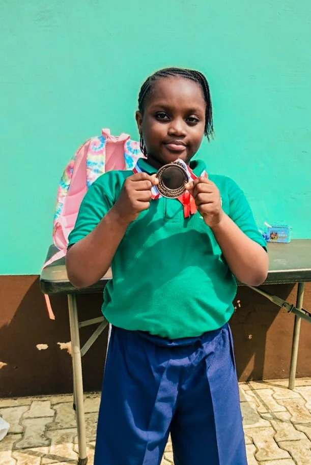 Building exceptional students at Fels Ideal Academy. A young girl wearing a green shirt and blue shorts holds up a silver medal from a Math competition.