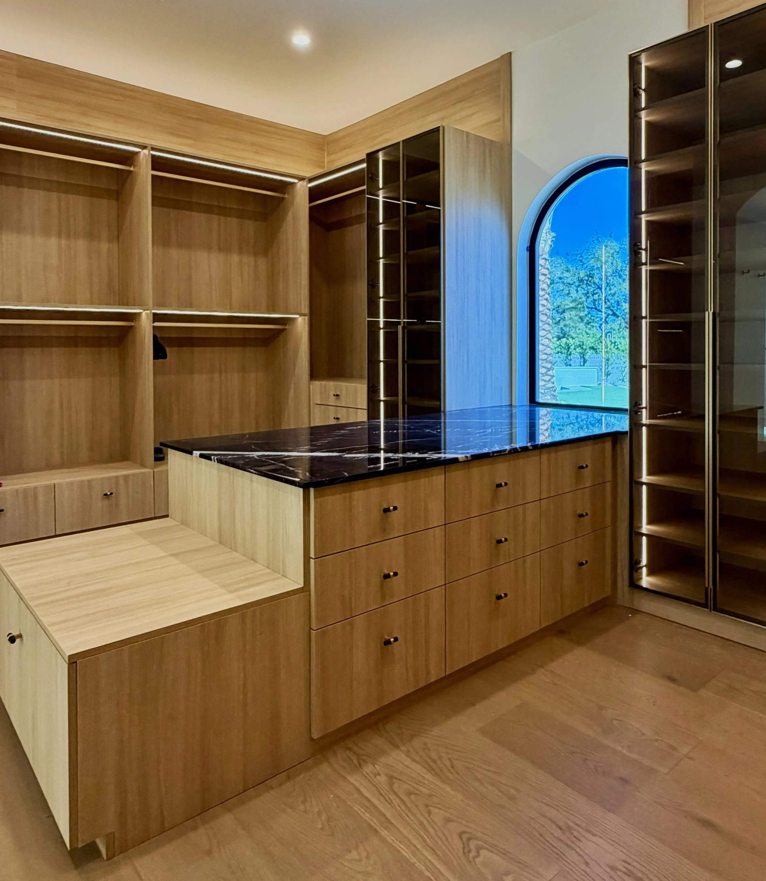 walk in master closet, seating on island end, stone countertop, drawers on island, full high gold glass doors, integrated lighting in all areas, including above closet cabinets
