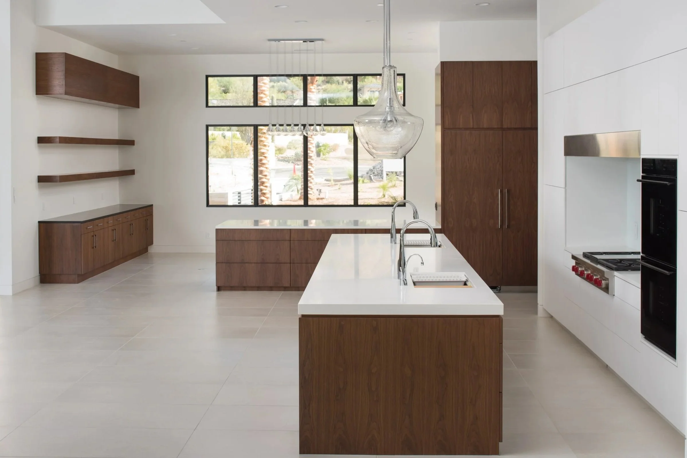 Modern kitchen with white countertops, wooden cabinets, and a large island. Black-framed windows in the background let in natural light. Open space with minimal decor.