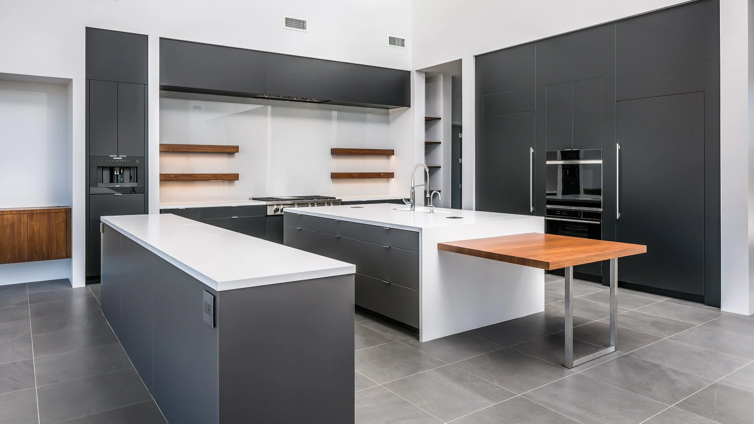 Modern kitchen with black and white cabinets, wooden shelves, gray tiled floor, and a large kitchen island with a built-in sink.