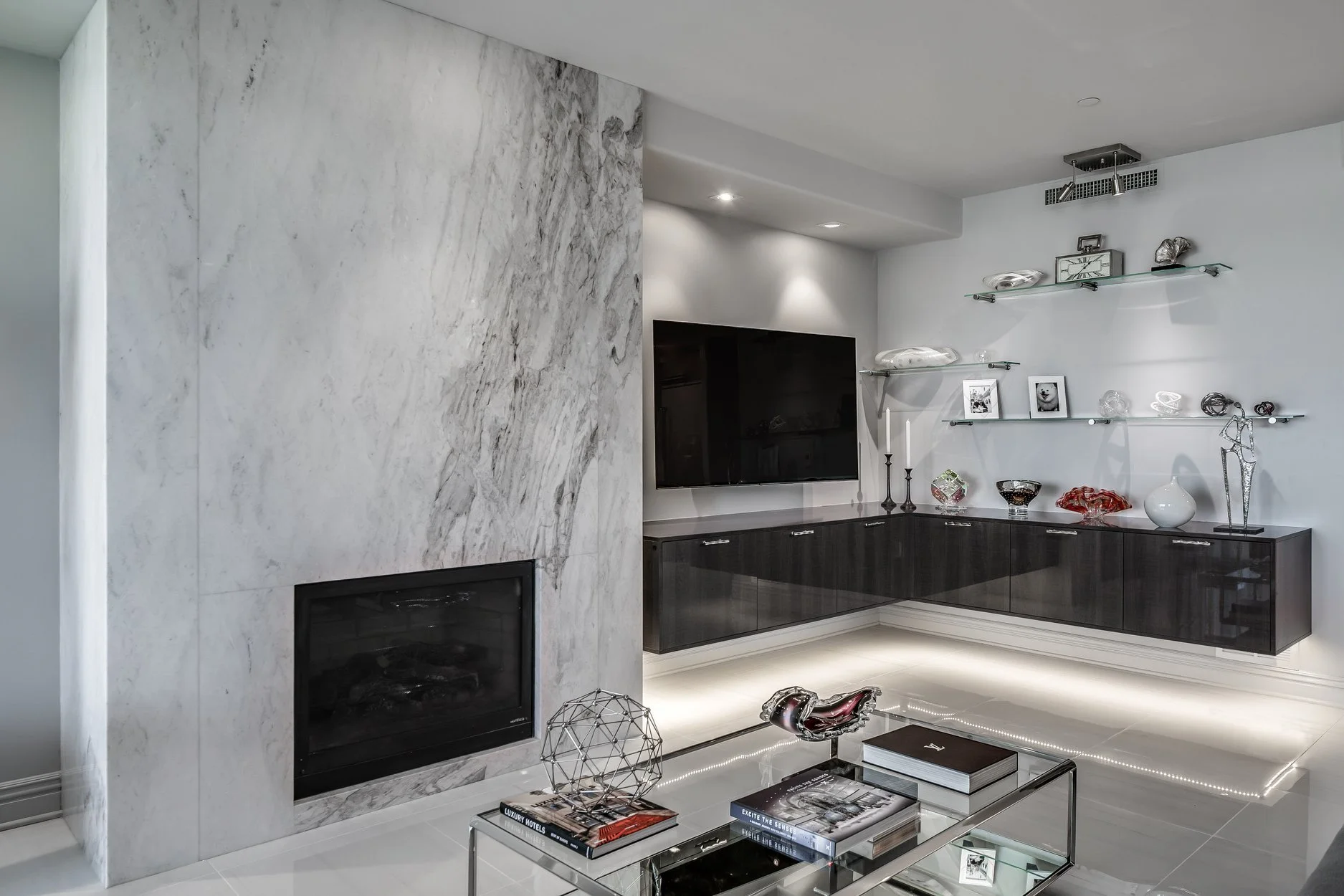 Modern living room featuring a gray marble fireplace, a black wall-mounted TV, floating glass and metal shelves with decorative items, and a glass coffee table with books and sculptures.