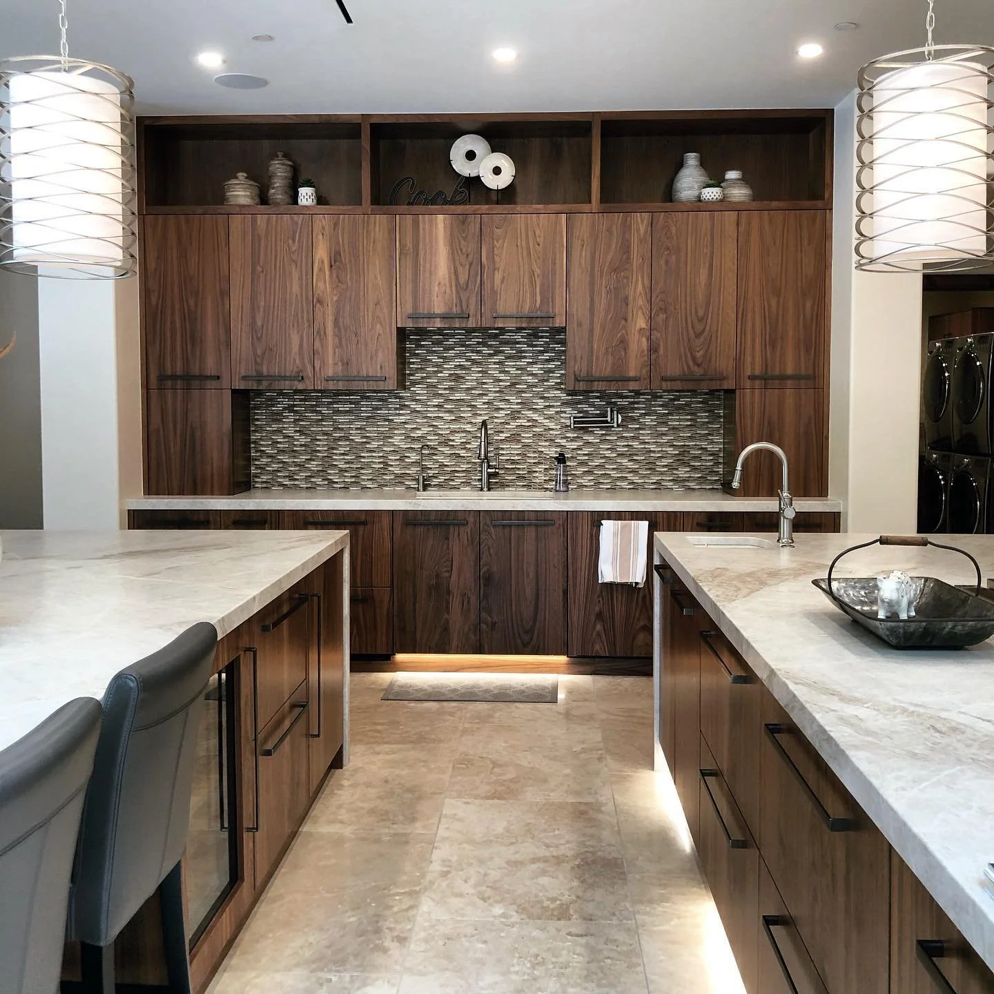 Modern kitchen with dark wood cabinets, a beige stone countertop, and a tiled backsplash with horizontal mosaic pattern, illuminated under the cabinets. There are hanging pendant lights and a built-in open shelf with decorative items.