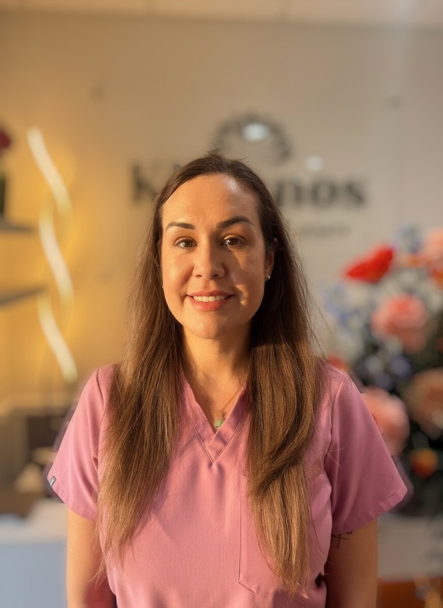 A woman with long brown hair wearing pink scrubs, smiling indoors with a blurred background including flowers and a sign.