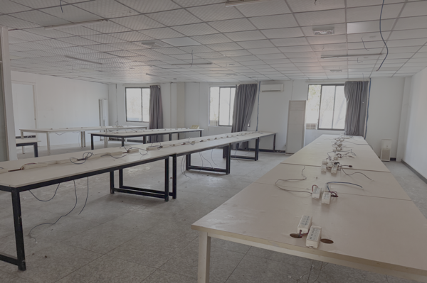 A room in scam compound office building with three windows. It's empty except for and five long tables with power strips and computer cables.