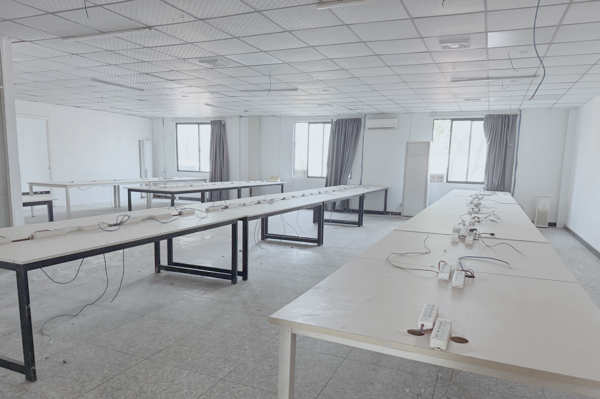 A room in an abandoned building. There are four rows of long tables with closely spaced power strips and wiring. The walls and floor are white.