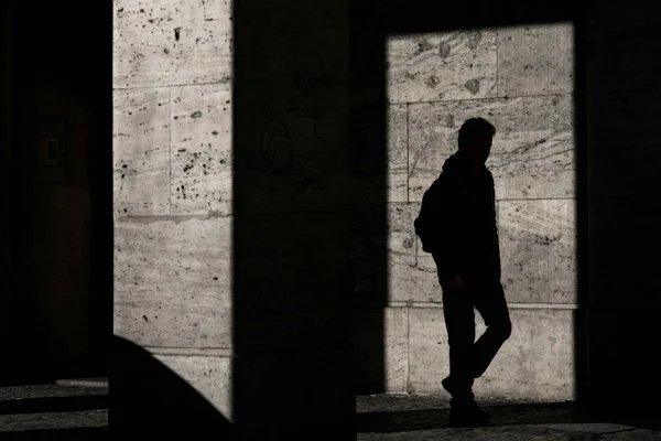 A silhouette of a person walking in a doorway sized section of light near a shadowed concrete wall.