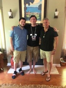Three men posing in front of a painting of a beach. One has a blue shirt, the others have black shirts.