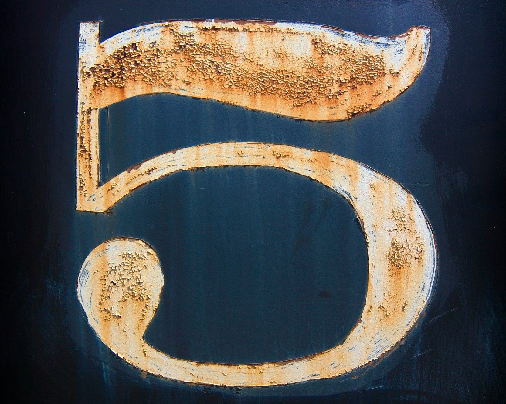 The numeral five painted on a dark blue background. The number is white paint that with rust from the metal beneath it.