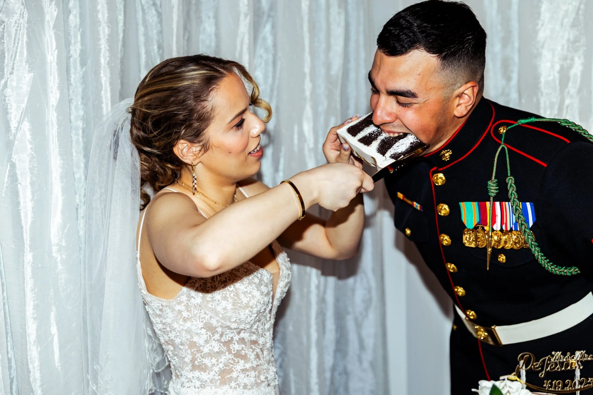 A woman in a wedding dress holding a cake slicing it for a man in a military uniform, who is biting into a piece of cake.