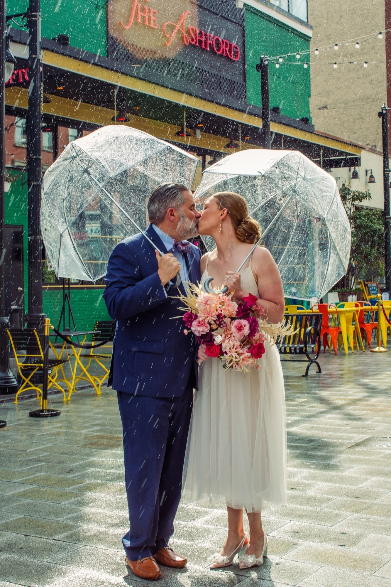 A bride and groom sharing a kiss under umbrellas in the rain outside a venue called The Ashford, with colorful chairs and string lights in the background.