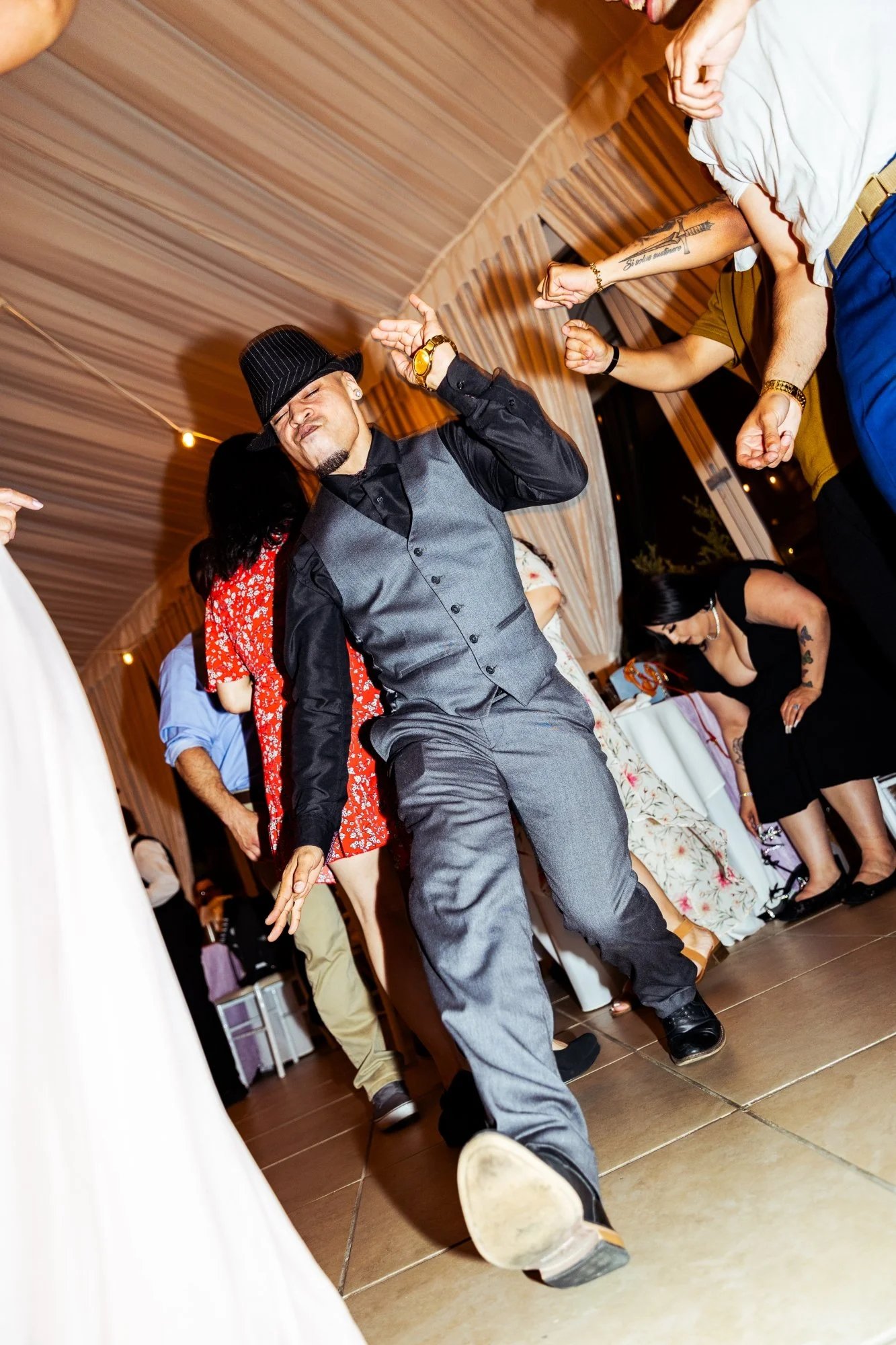A man wearing a gray suit, black shirt, black hat, and gold watch dancing at a party with other people around him. The setting appears to be indoors with draped curtains and string lights.