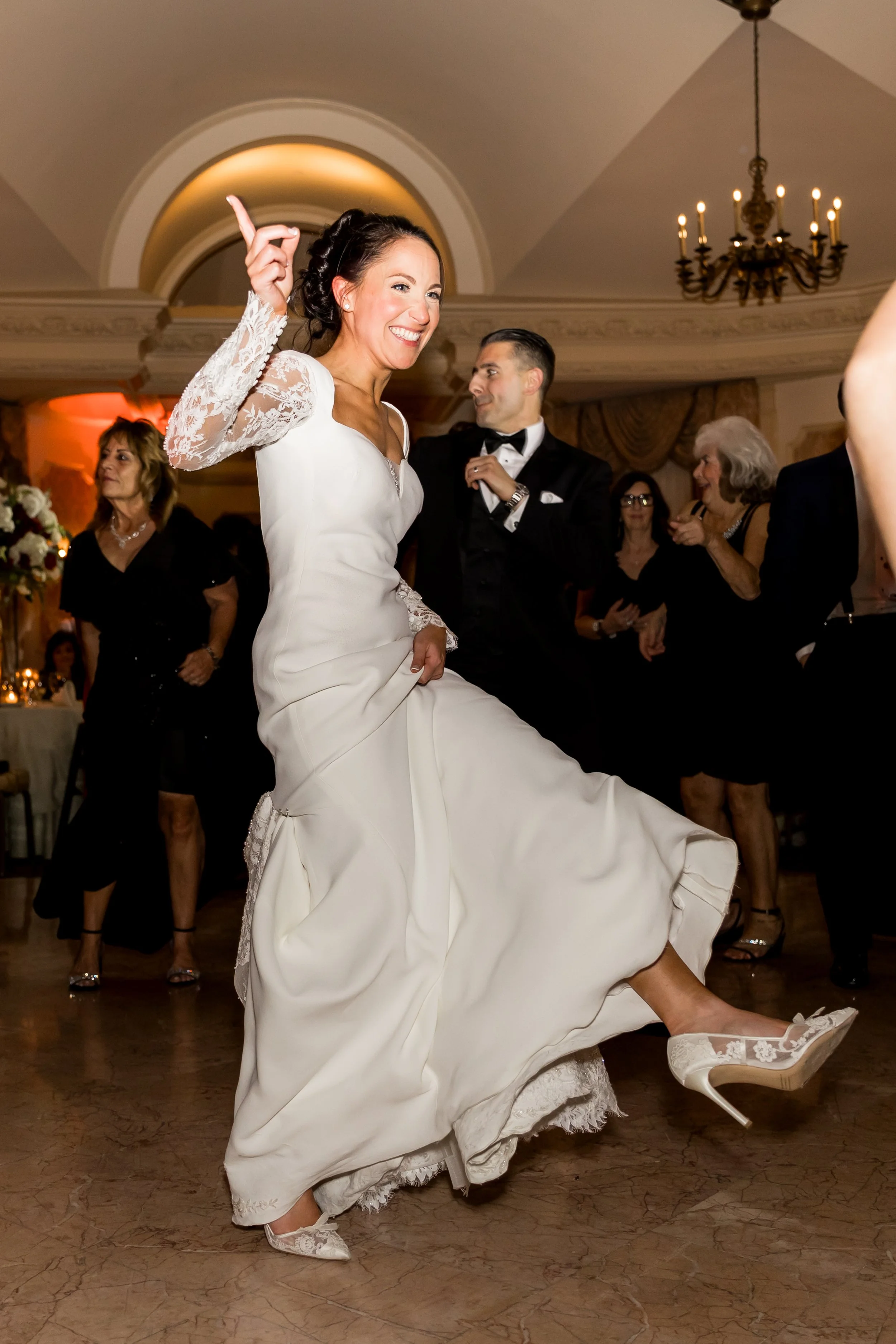 A bride in a white wedding dress dancing and lifting her skirt, smiling, at her wedding reception, surrounded by guests in an elegant indoor setting.