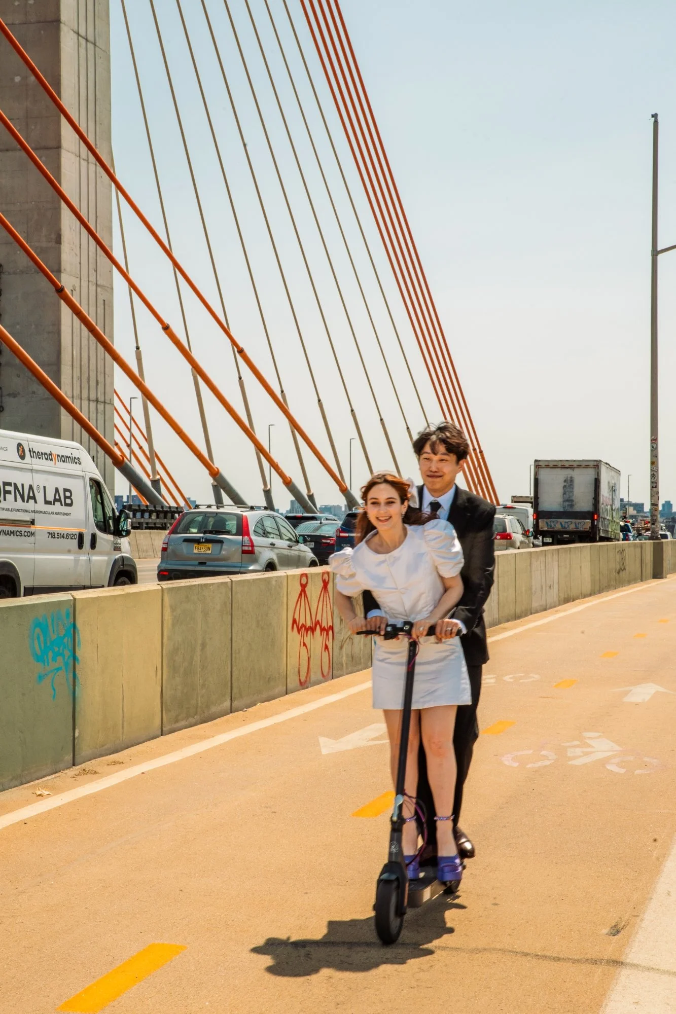 A man and woman riding an electric scooter together on a designated bike lane under a bridge with red cables, in an urban setting with cars and trucks in the background, and graffiti on a concrete wall.