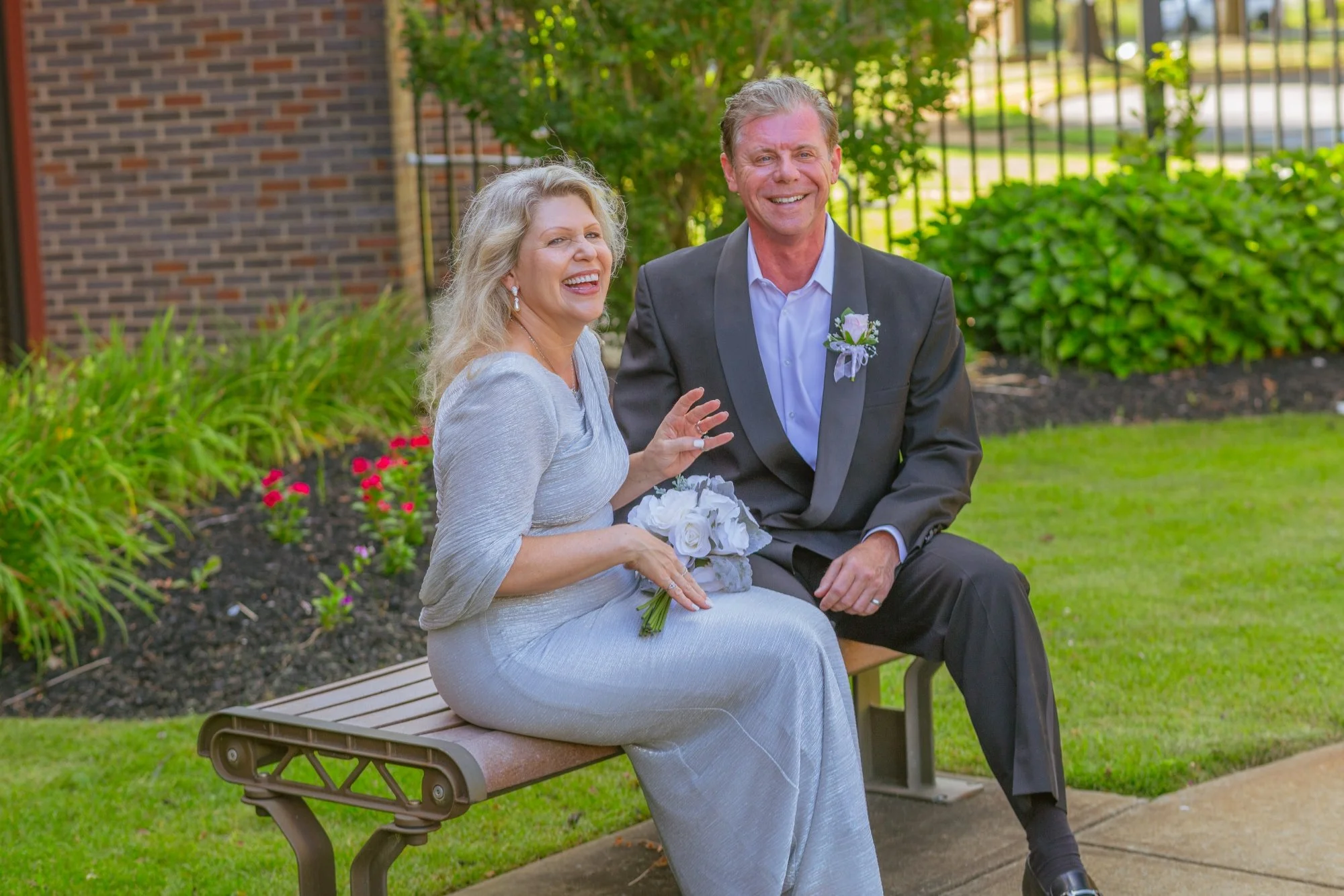 A smiling couple sitting on a park bench outdoors, with the woman dressed in a light gray gown holding a bouquet of white flowers, and the man in a black suit with a boutonniere, enjoying a sunny day near a brick building and lush green bushes.