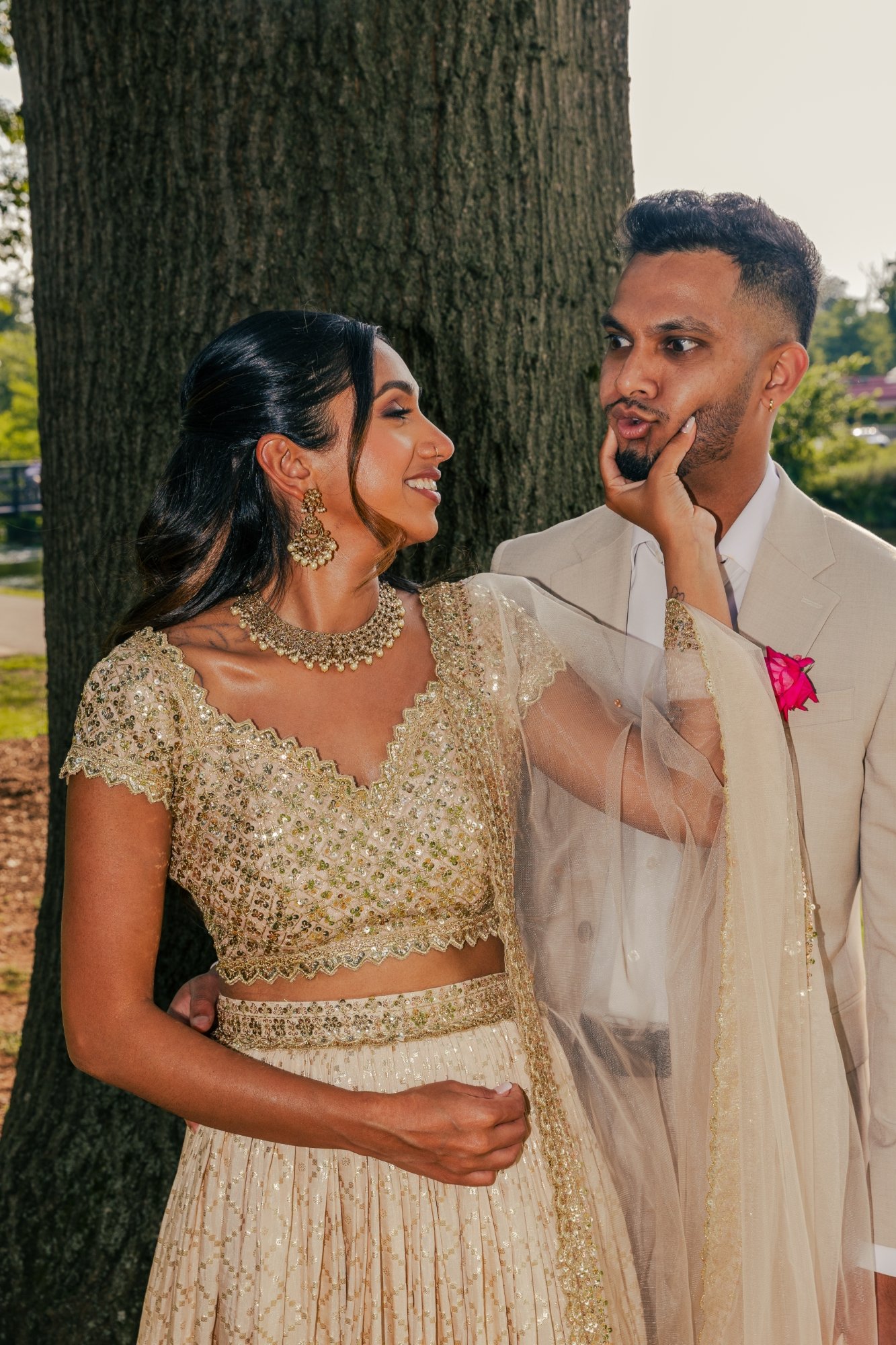 A woman in a gold traditional dress playfully touches a man's chin in an outdoor setting near a tree, both smiling and dressed in elegant attire.