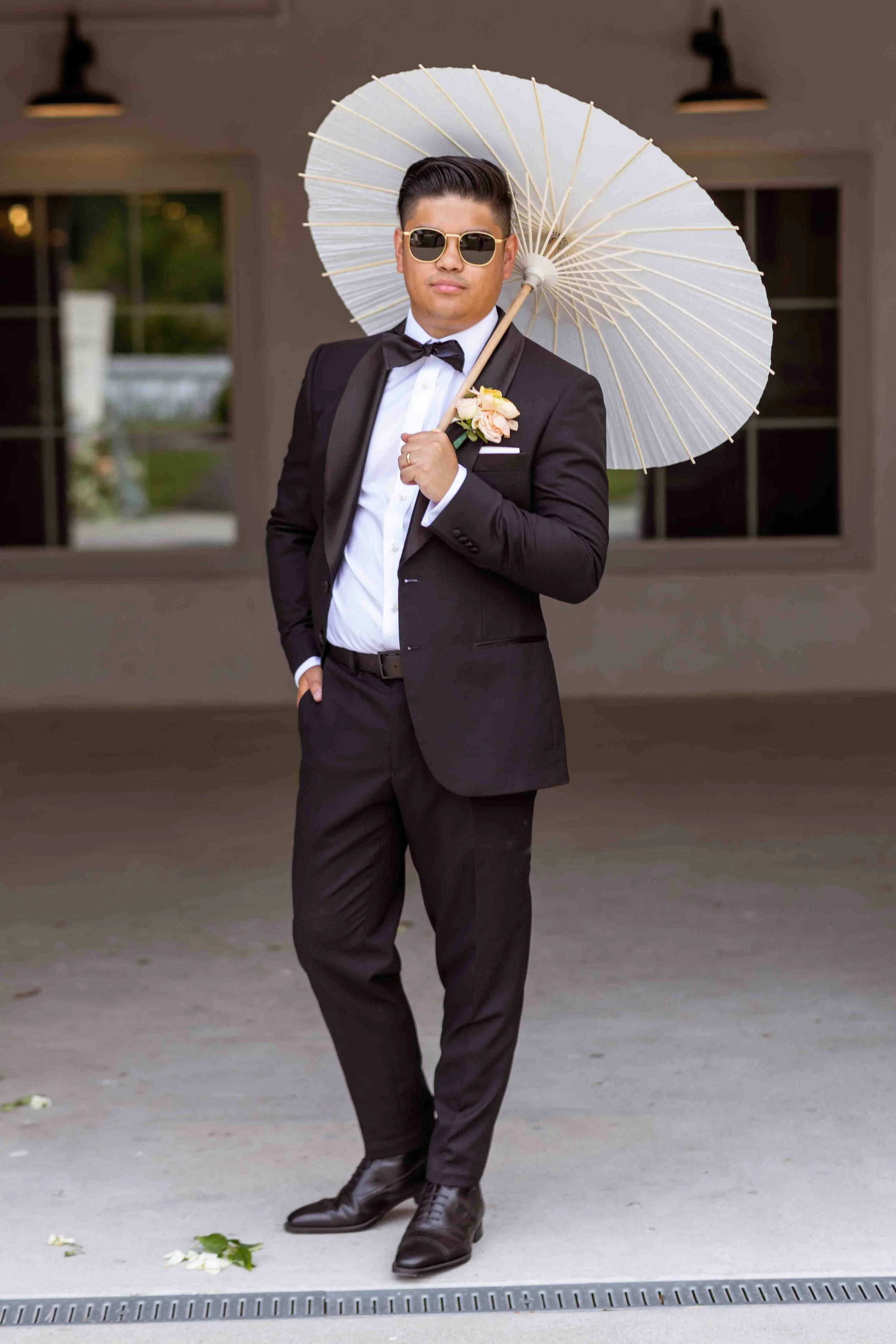 A man in a tuxedo holding a white parasol, wearing sunglasses, standing outdoors with a building in the background.
