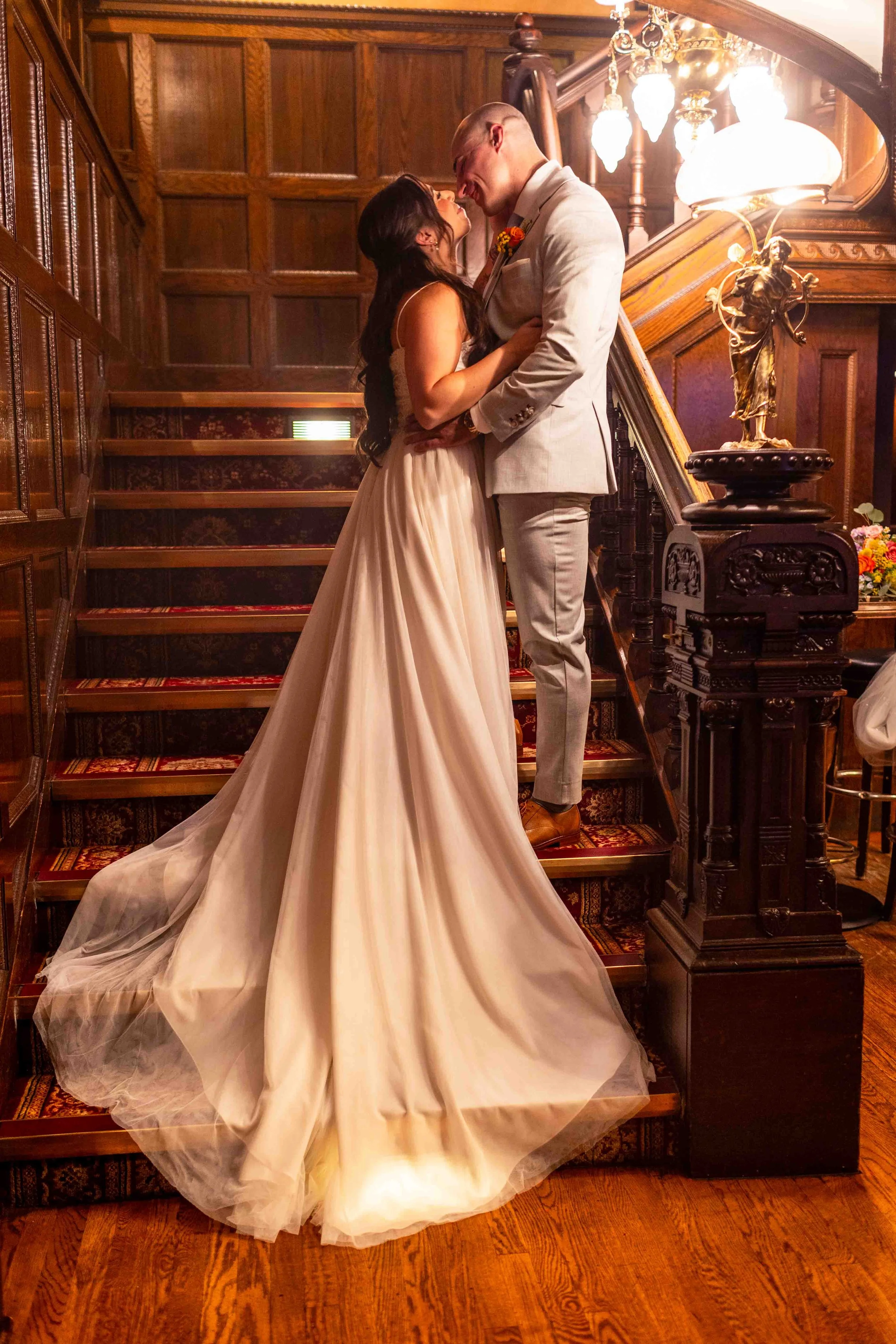 A bride and groom sharing a kiss on a staircase inside a wooden-paneled room, with warm lighting and decorative details.