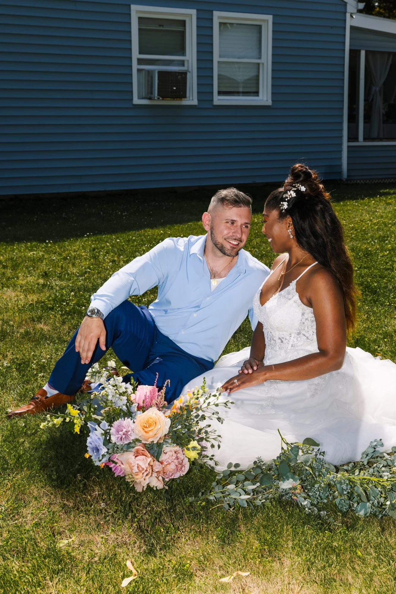 A newlywed couple sitting on the grass in front of a blue house. The groom is dressed in a light blue shirt and blue pants, while the bride is wearing a white wedding dress with lace details. They are smiling and looking at each other. There is a colorful bouquet of flowers in front of them and greenery arranged on the ground.
