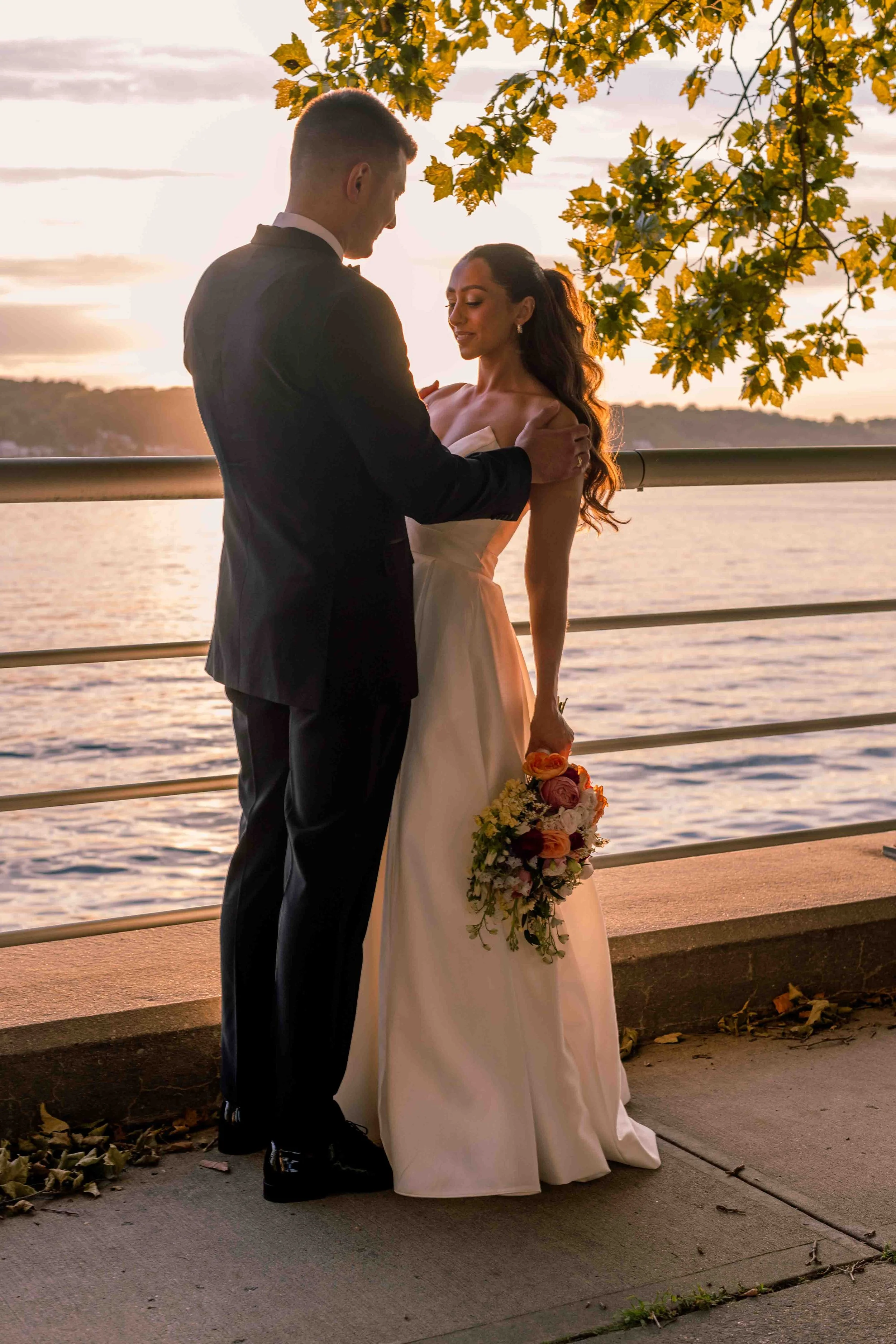 A bride and groom standing near a railing by a body of water during sunset, sharing an intimate moment. The bride is holding a bouquet of flowers, wearing a white gown, and the groom is in a dark suit.