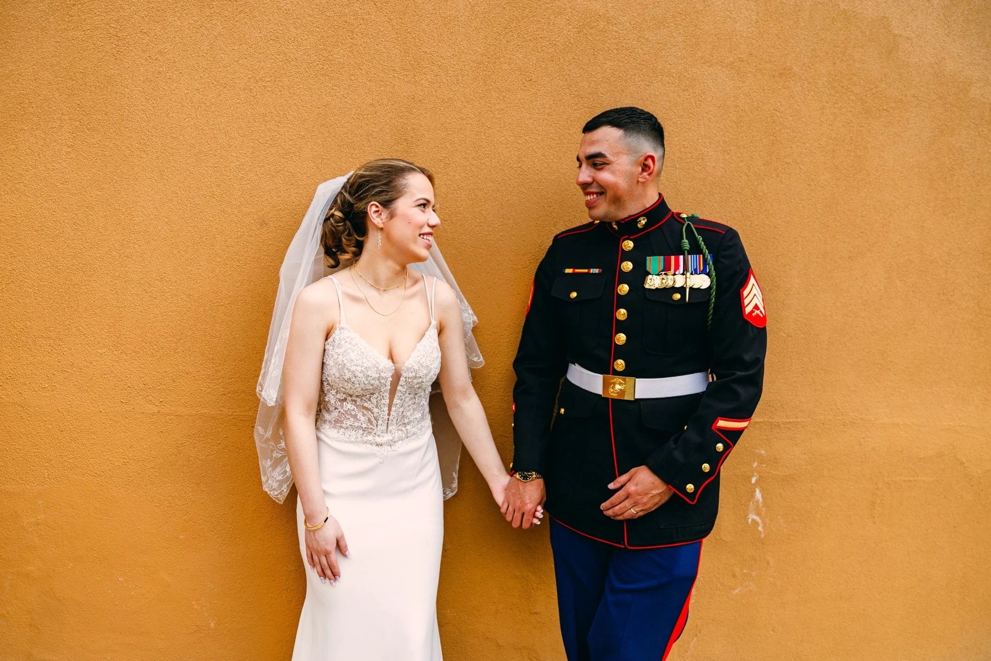 A bride and a groom in military uniform holding hands and smiling at each other against a tan wall.